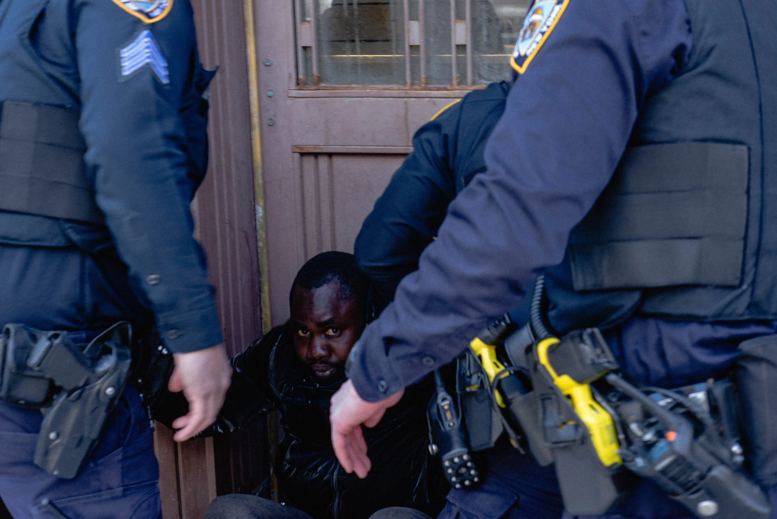 Two police officers are restraining a man on the floor near a wooden door with vertical metal bars. The man appears to be handcuffed or restrained and is looking at the camera with a serious expression.