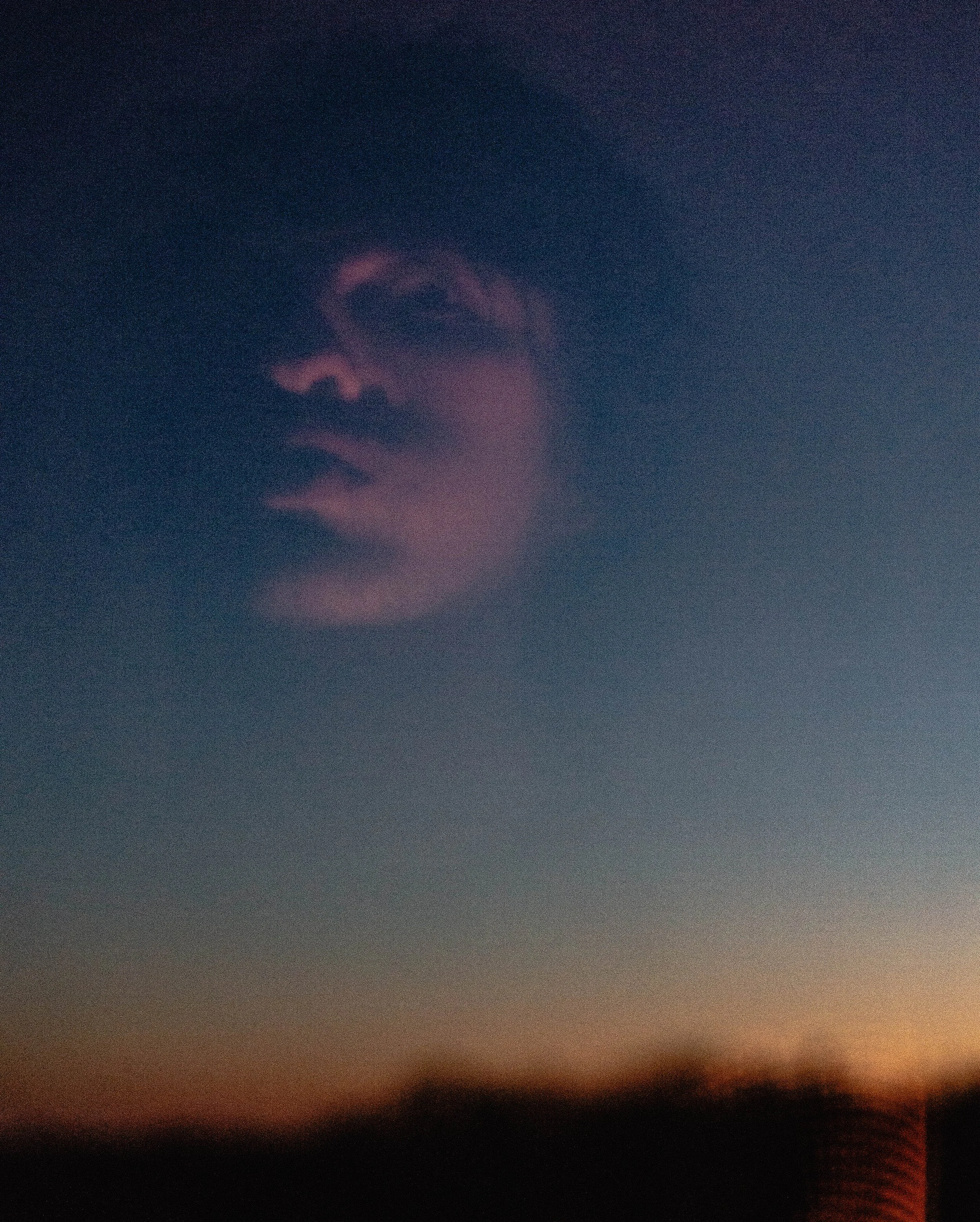 A reflection of a person's face with glasses is seen in a mirror or glass surface against a blurred sunset or twilight sky.