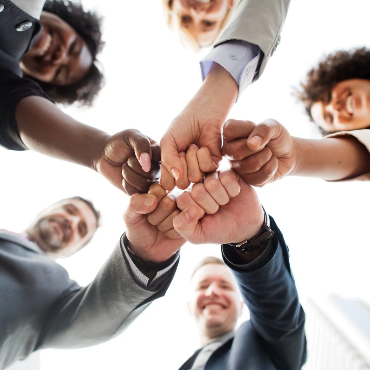 Group of diverse professionals in business attire putting their hands together in a circle, viewed from below, smiling and symbolizing teamwork and collaboration.
