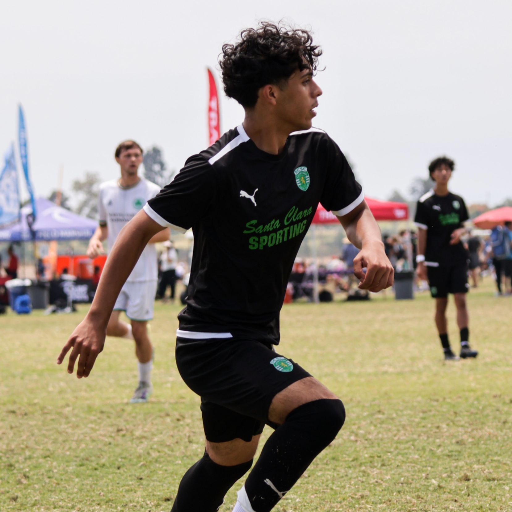 A young soccer player in a black uniform running on a grass field during a match, with other players and tents in the background.
