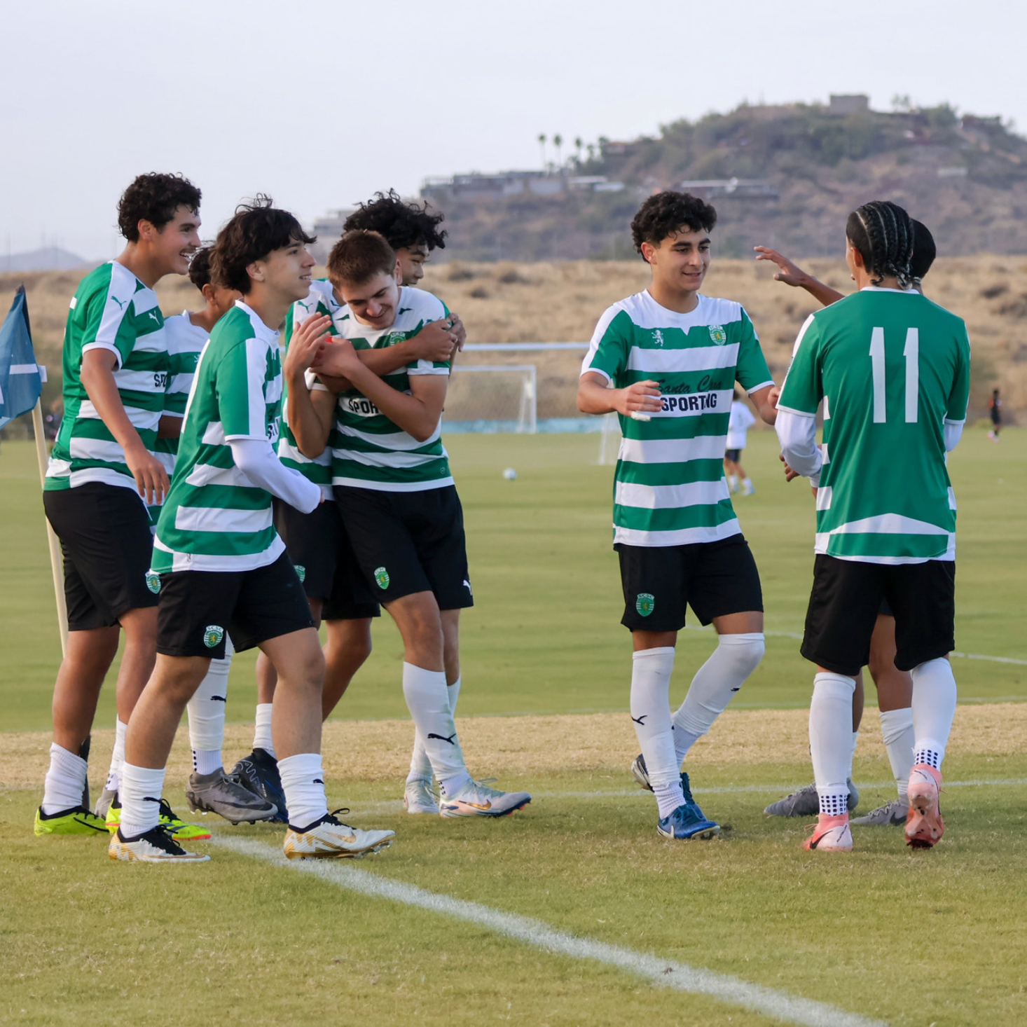 Group of young soccer players in green and white uniforms celebrating on the field, some smiling and embracing each other.