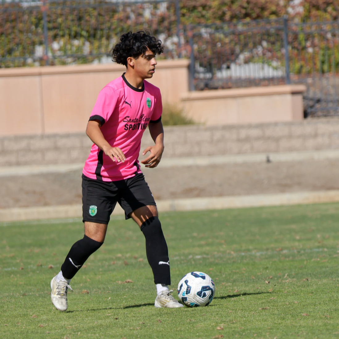 A young man playing soccer outdoors, wearing a pink jersey and black shorts, with a soccer ball at his feet.