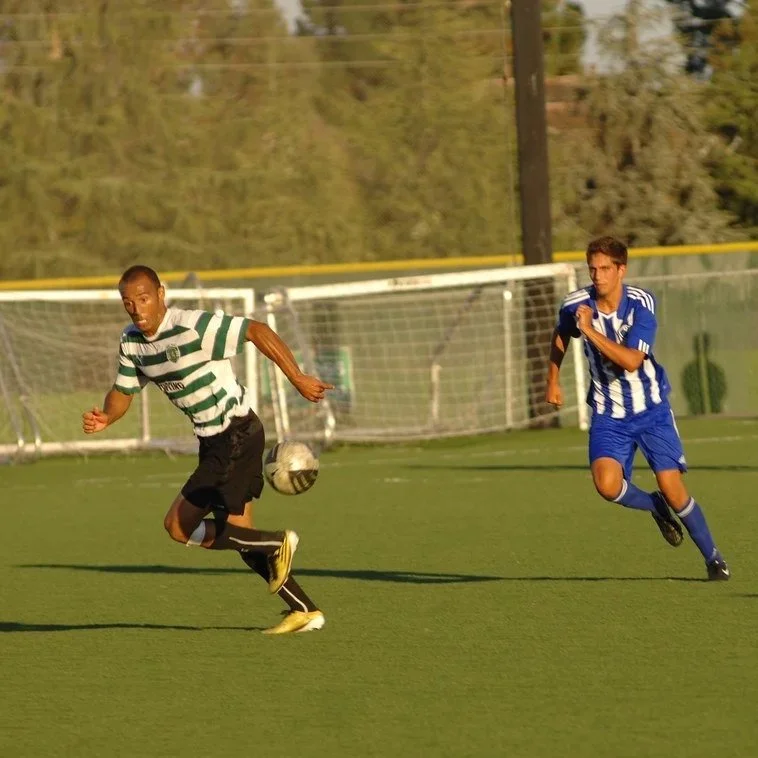 Two young male soccer players in green and white striped and blue and white striped jerseys running after a soccer ball on a grassy field during daytime.