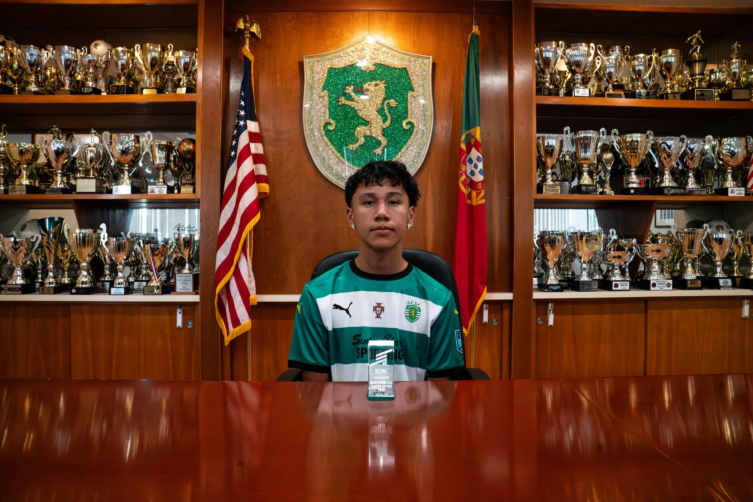 A young person sitting at a conference table in front of a wall filled with numerous trophies and awards. Behind them are the flags of the United States and Portugal, as well as a crest with a lion on a shield.