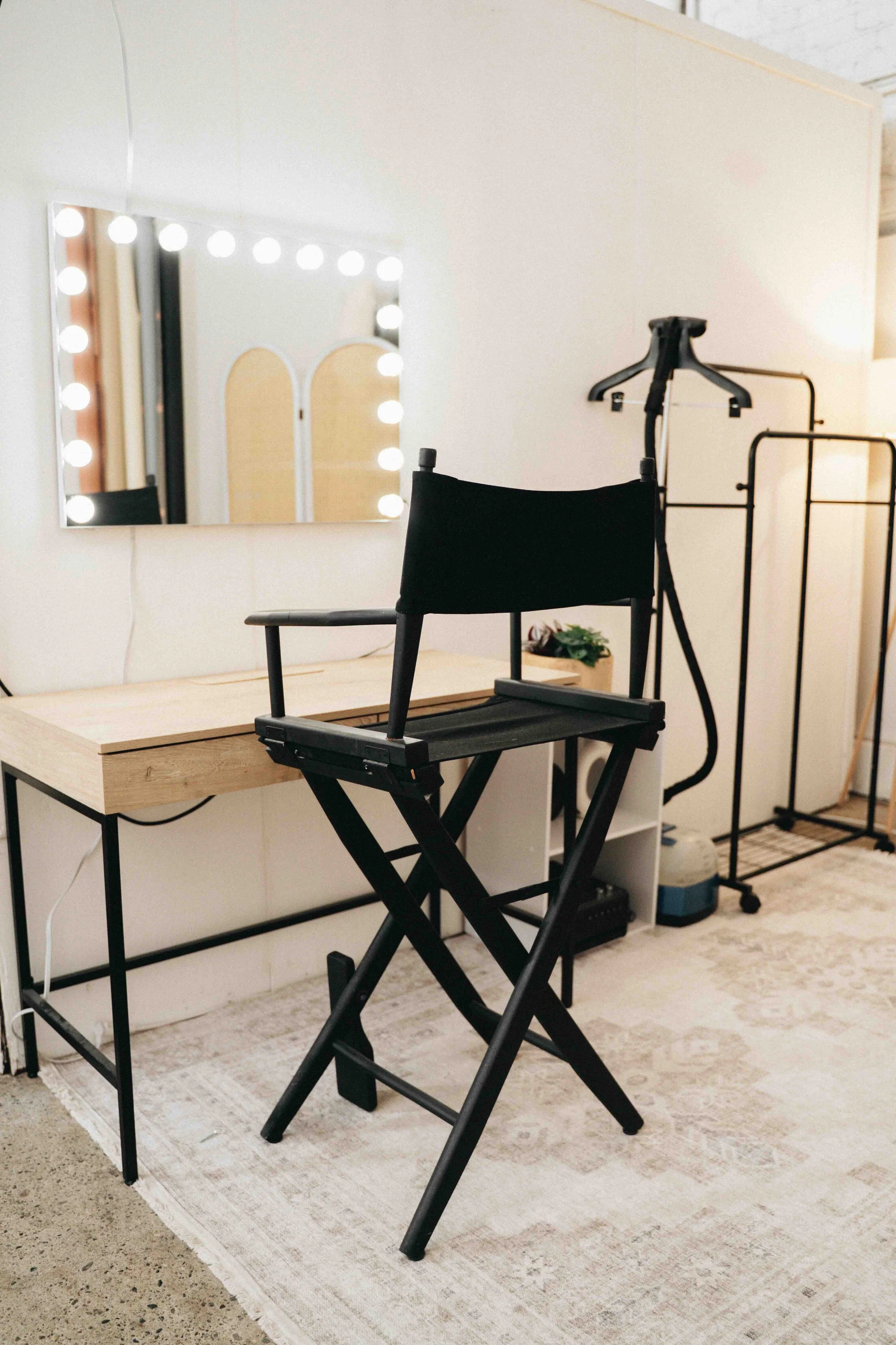 A makeup vanity area with a mirror surrounded by lights, two black director's chairs, a wooden table, and a clothing rack in a white brick-industrial space.