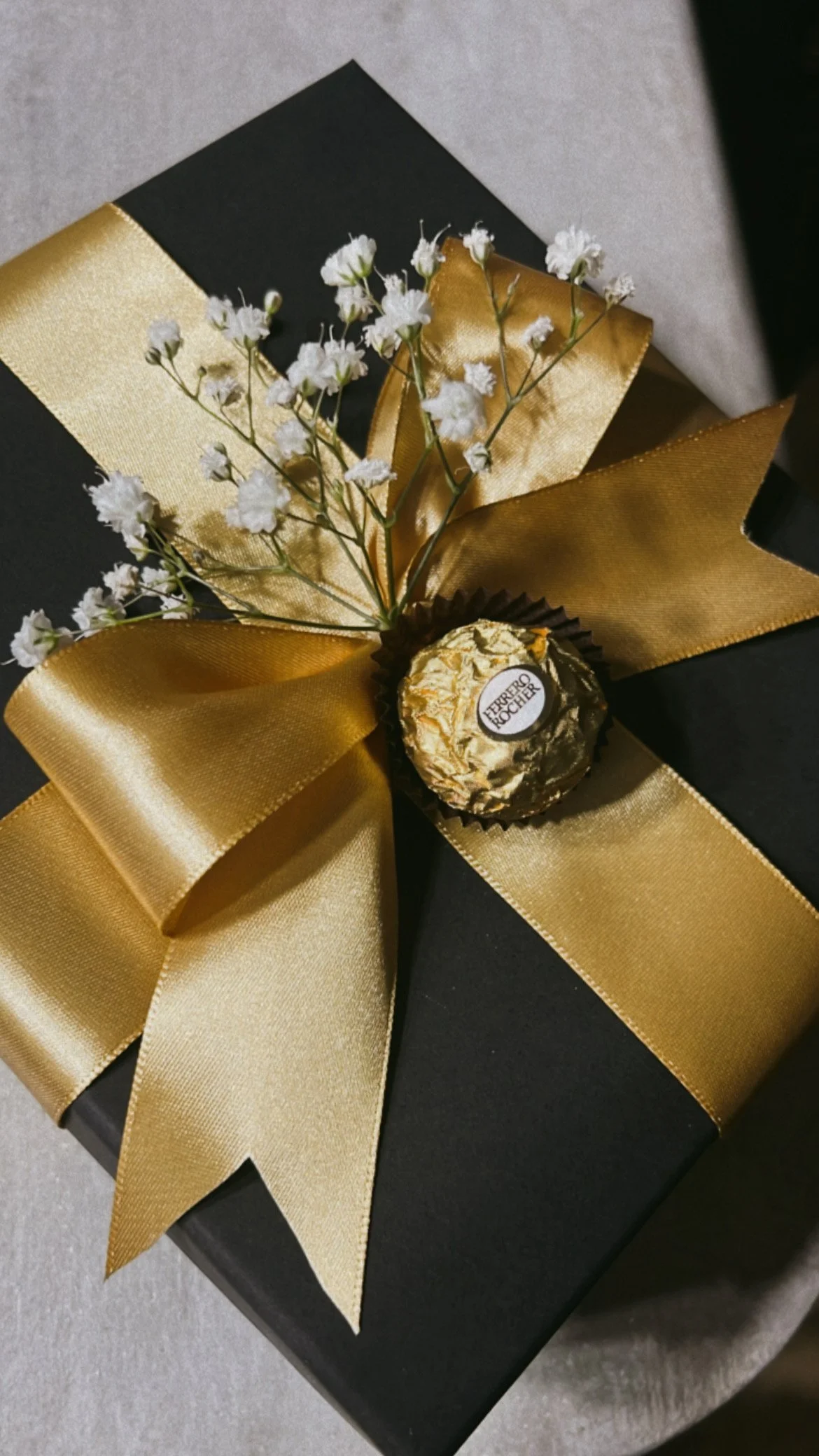 A black gift box decorated with a large gold satin ribbon, a small sprig of white baby's breath flowers, and a gold Ferrero Rocher chocolate on top.