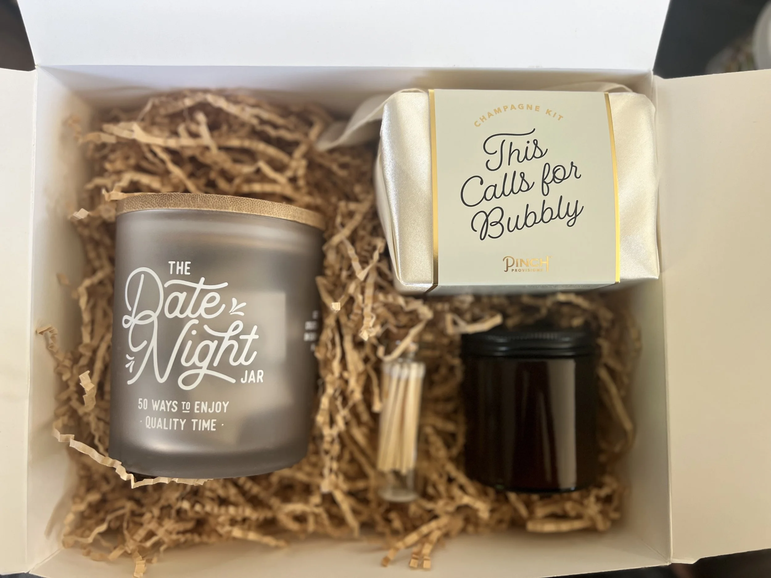 Gift box containing a candle, a small bottle, a box labeled 'This Calls for Bubbly,' and a small jar, all nestled in shredded paper.
