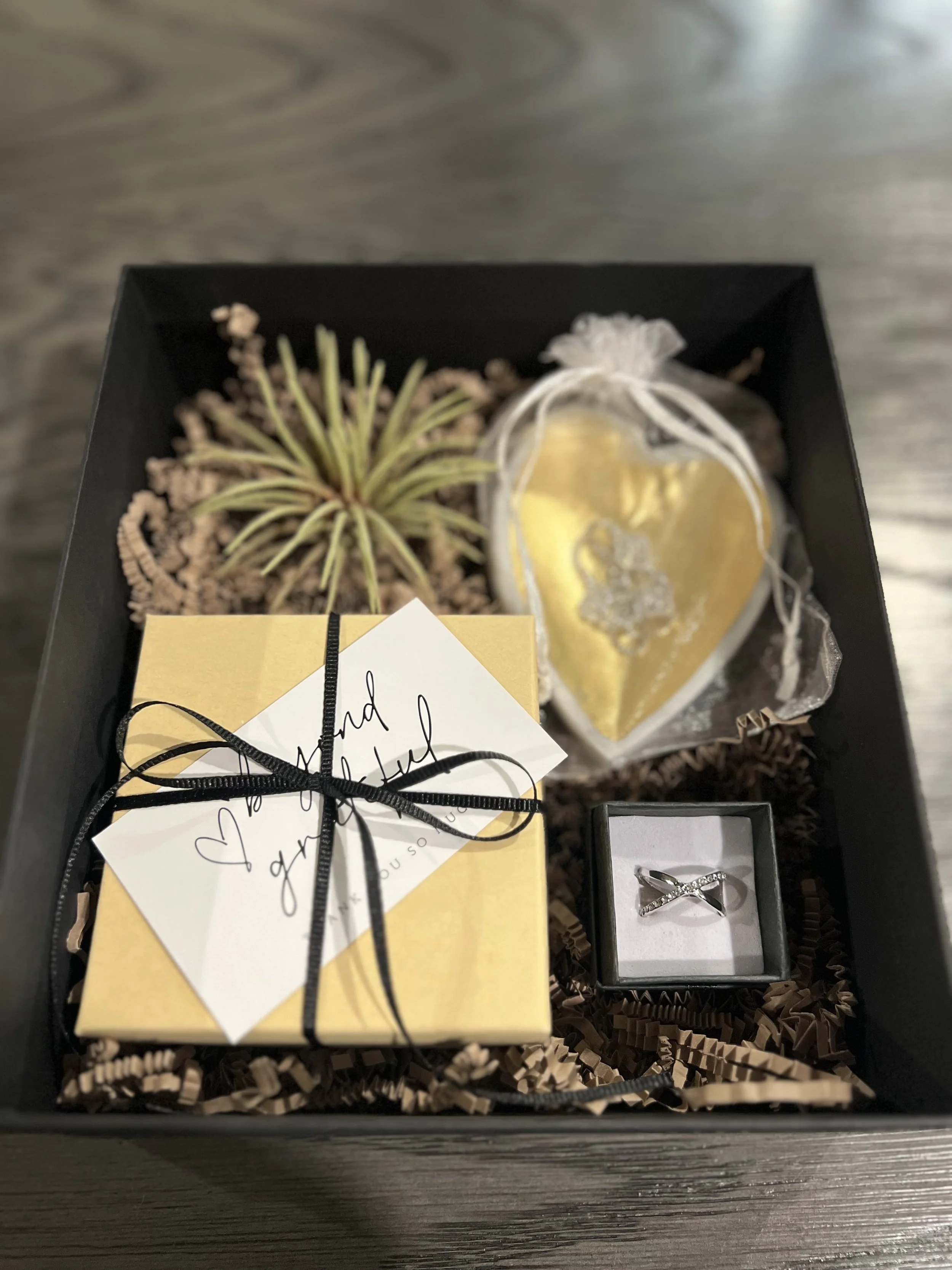 A gift box containing a small potted plant, a gold heart-shaped ornament, a yellow box tied with a black ribbon and a handwritten note, and a ring in a black box.