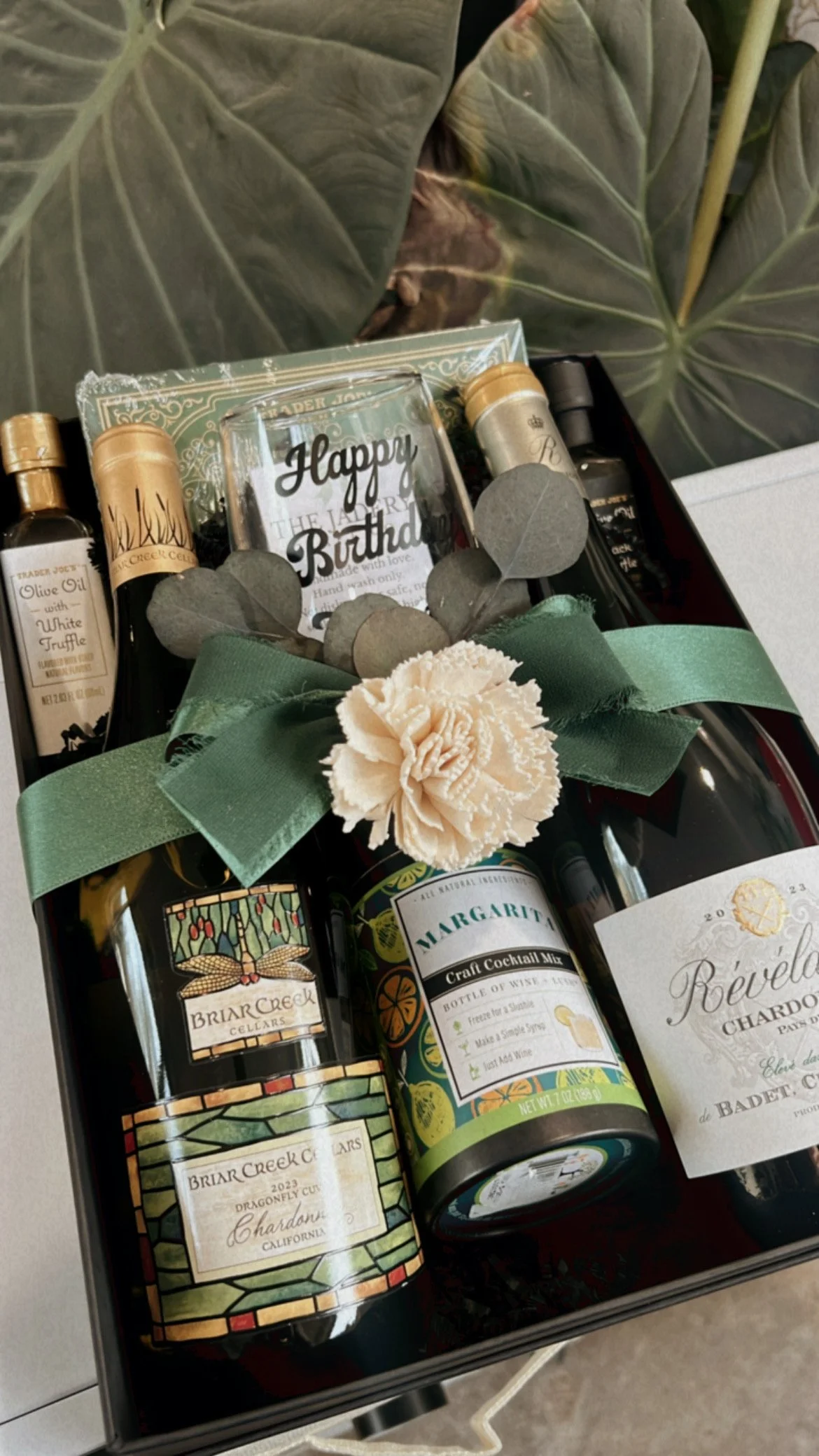 A gift box with two bottles of wine, a small bottle of cocktail mix, a glass with 'Happy Birthday' written on it, and a decorative flower with greenery, all tied with a green ribbon. The box is set against leafy background.