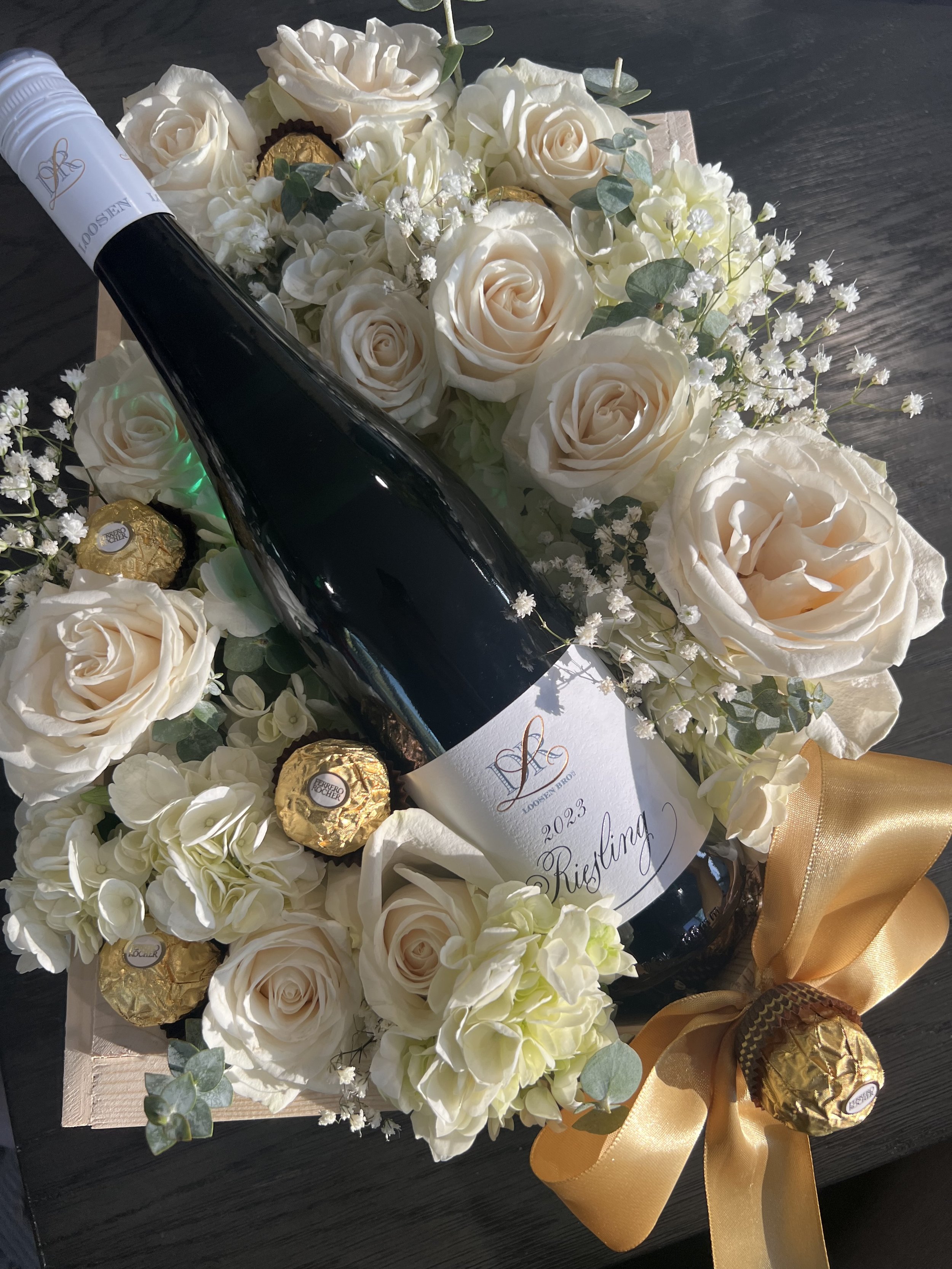 A bottle of sparkling wine with a white and gold label that says 2023, decorated with white roses, eucalyptus, baby's breath, and wrapped chocolates, all arranged in a floral display with a gold ribbon.