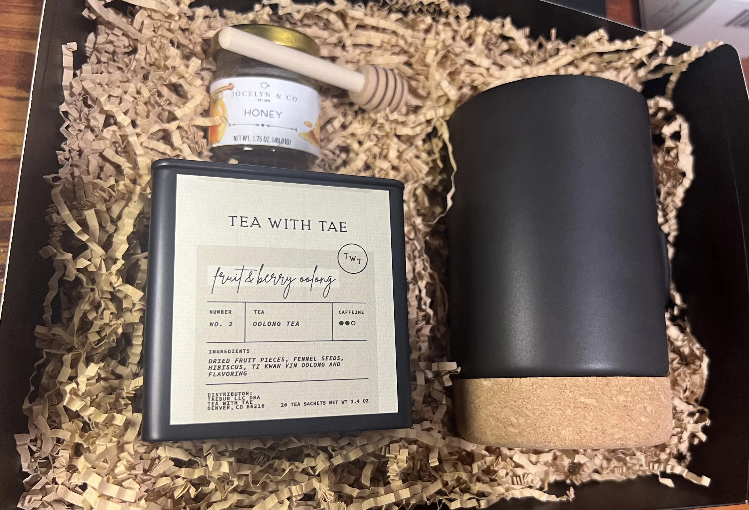 A gift box contains a jar of honey, a black cylindrical container with a cork base, a box of fruit and berry oolong tea, and a wooden honey dipper, all nestled in crumpled packing paper.