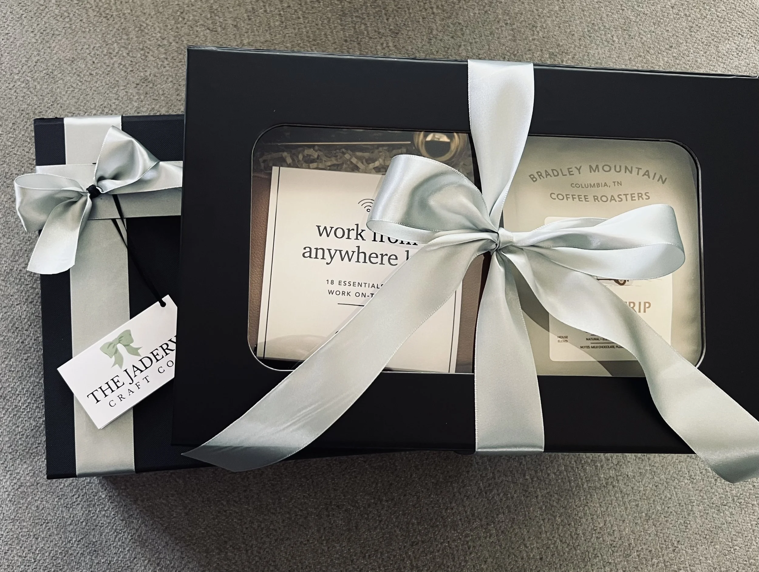 A black gift box with a clear window, decorated with a silver satin ribbon tied in a bow. Inside are a booklet titled 'work from anywhere' and a package from Bradley Mountain Coffee Roasters. There is a tagged card from The Jadeery Craft Co.