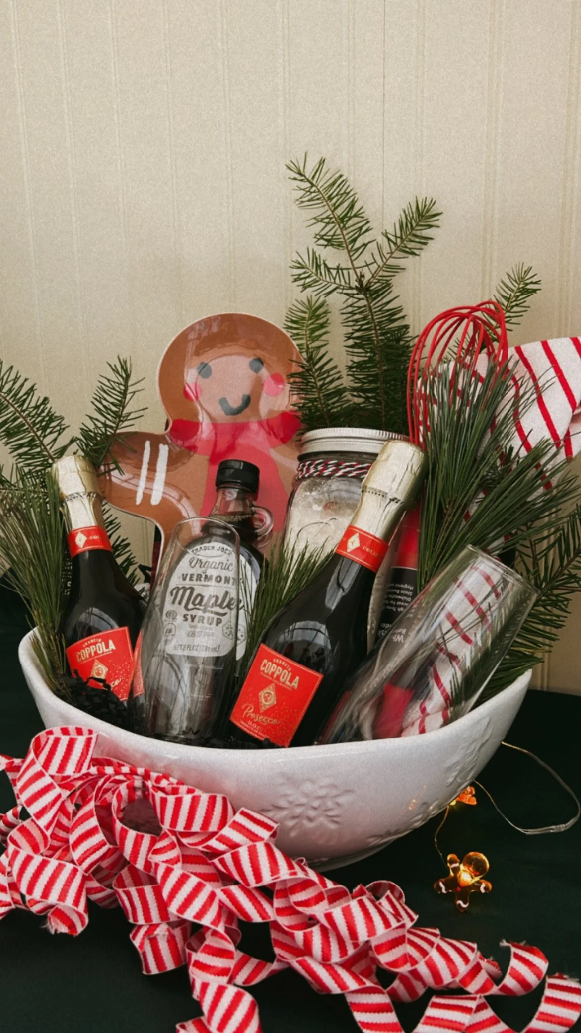 Christmas gift basket with small pine branches, bottled beverages, a gingerbread girl decoration, a jar, and red and white Christmas ribbons.