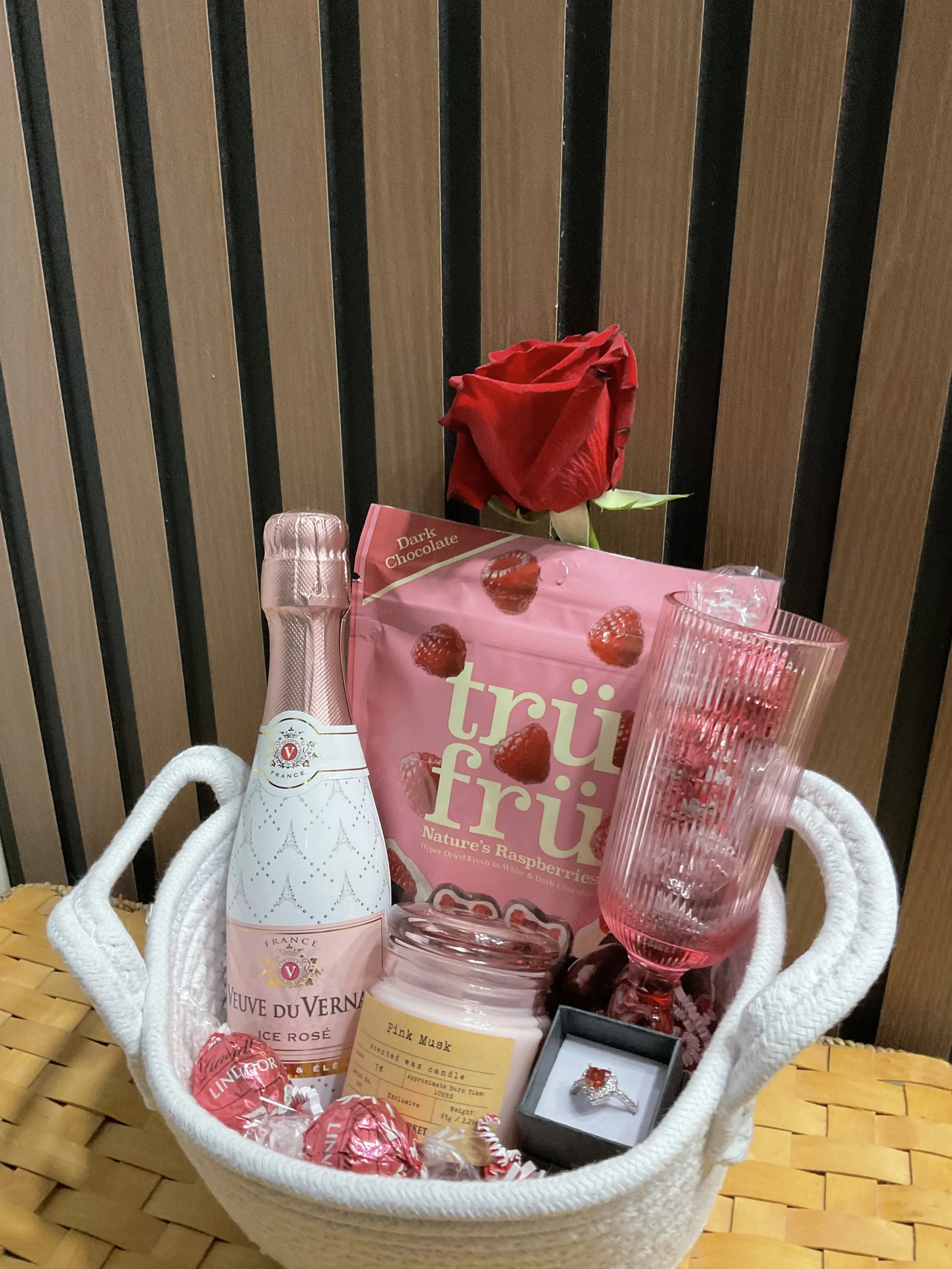 A gift basket containing a red rose, a pink box of dried raspberries, a small bottle of Veuve Clicquot Rosé champagne, pink candles, a glass with pink accents, Lindor chocolates, and a ring in a black box, set against a wooden background.