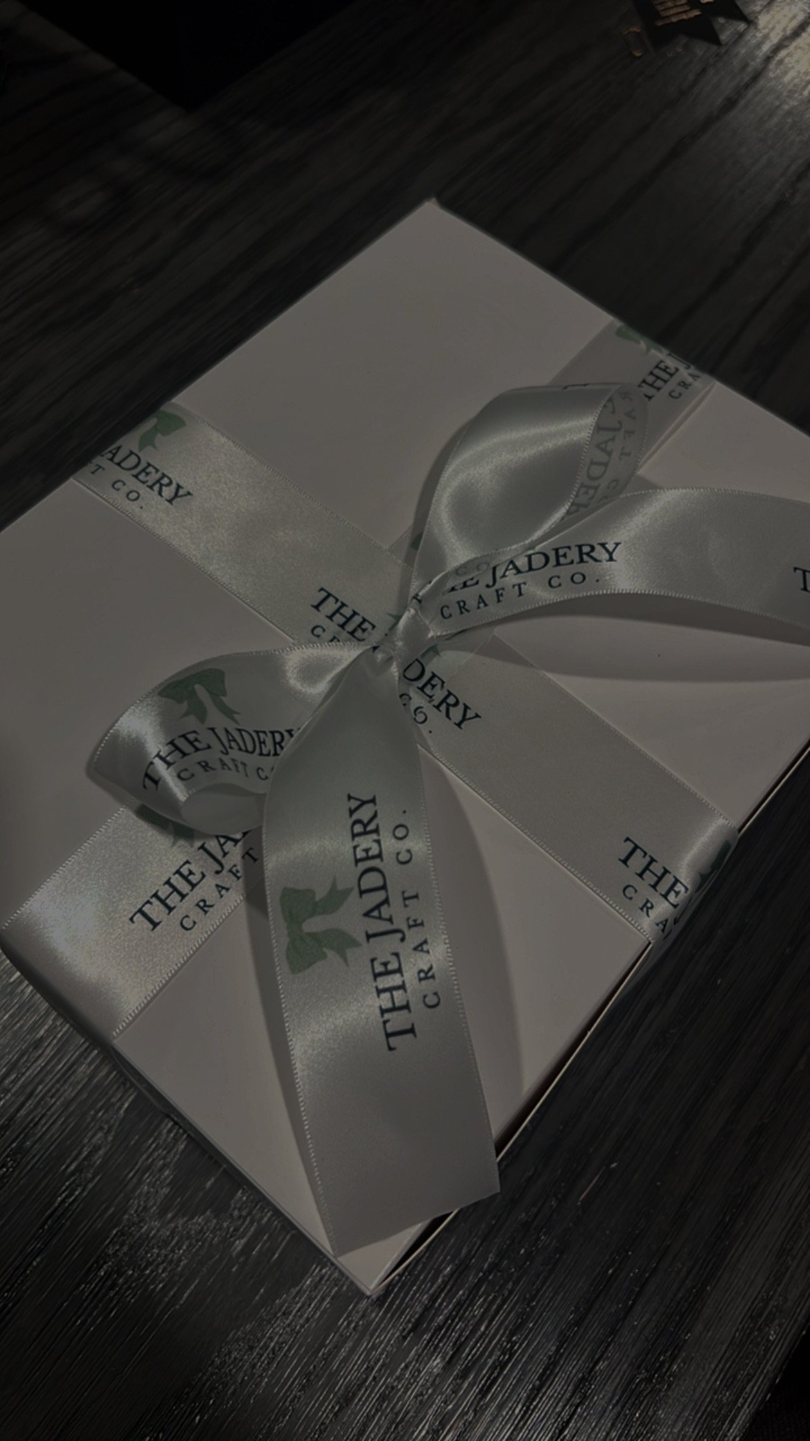 A white box with a white satin ribbon tied in a bow, labeled 'The Jader Craft Co.' placed on a dark wooden surface.