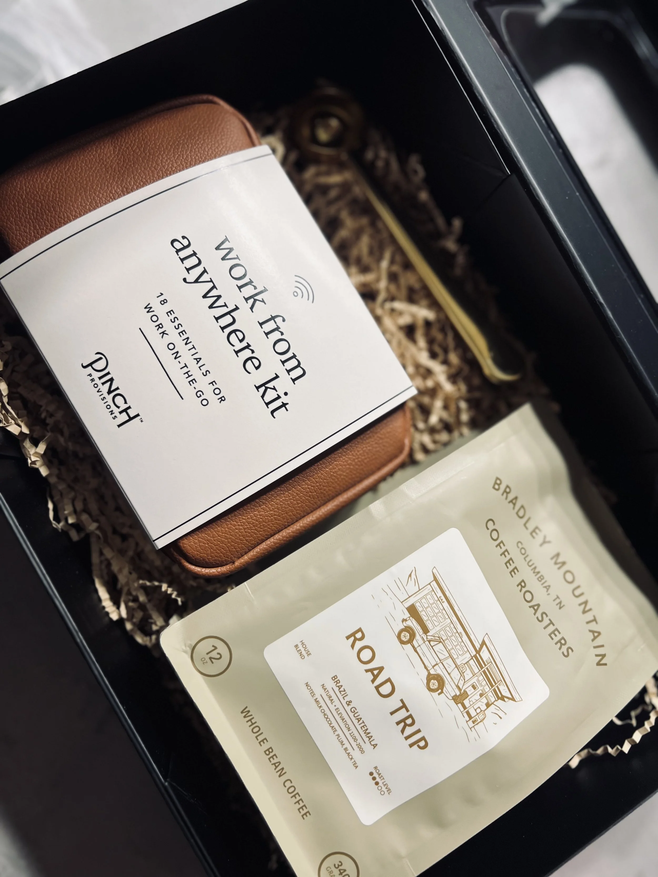 A gift box containing a brown leather wallet, a bag of Bradley Mountain coffee labeled 'Road Trip' from Columbia, Indiana, and a card with the message 'Work from anywhere kit, 18 essentials for work on the go'.