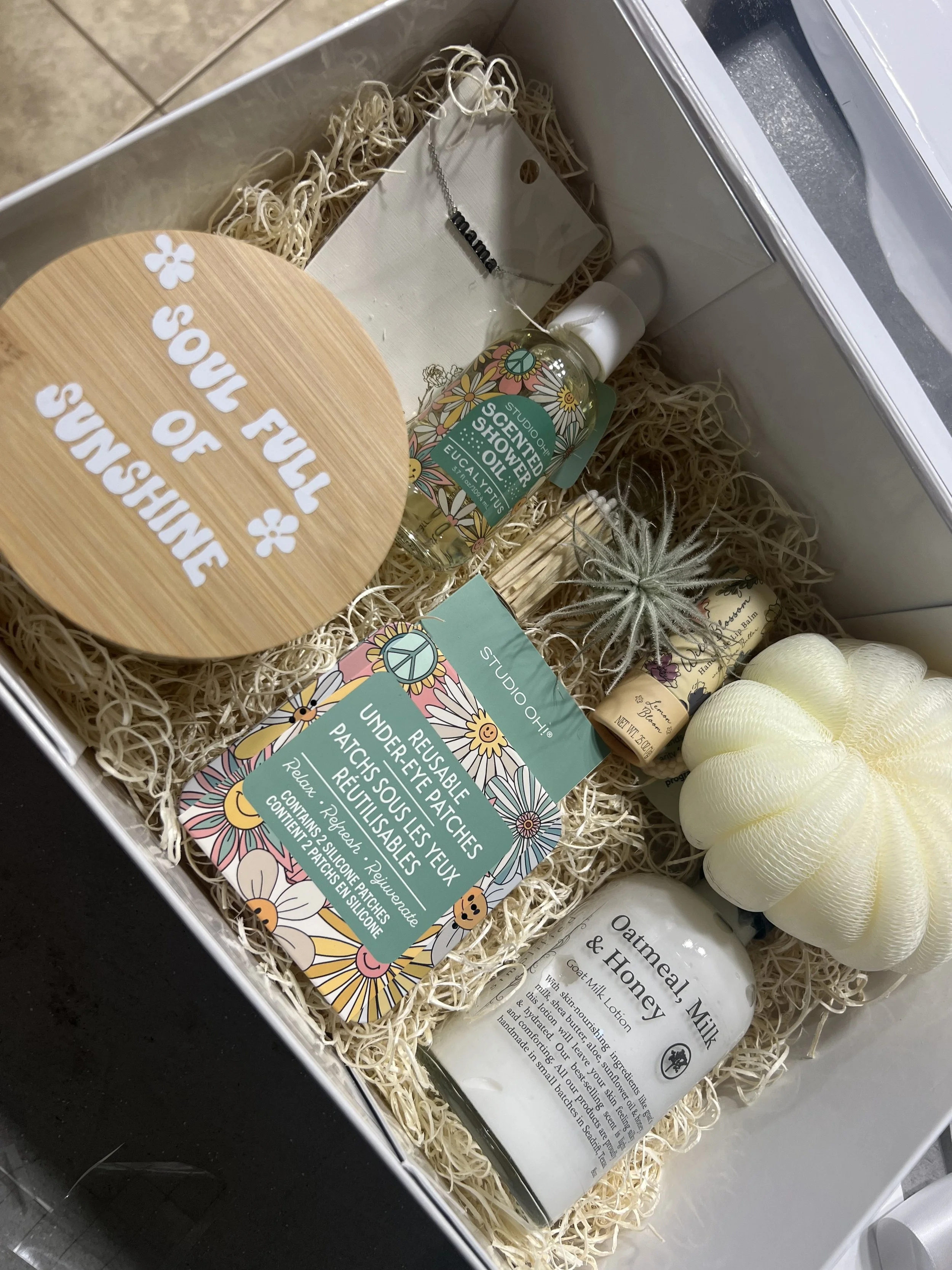 A gift box with natural filler material contains a bottle of oat and honey body lotion, a spray bottle of scented oil, a small candle, a decorative air plant, a bath bomb, some small rolled soap bars, and a round wooden sign that says 'You full of su