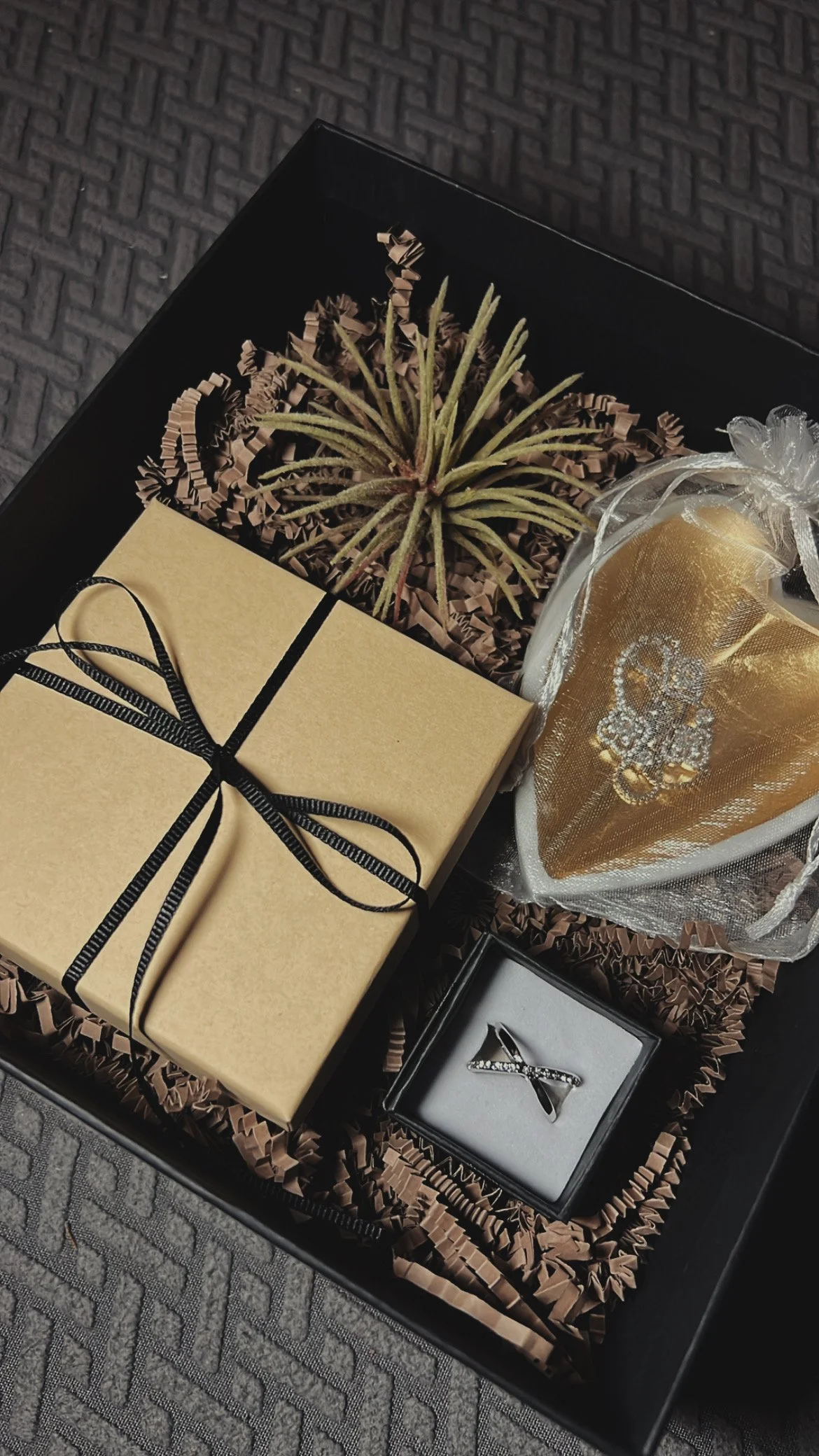 Gift box containing a potted plant, a gold heart-shaped item wrapped in sheer fabric, and a jewelry box with a silver ring, all arranged on shredded paper in a black tray.
