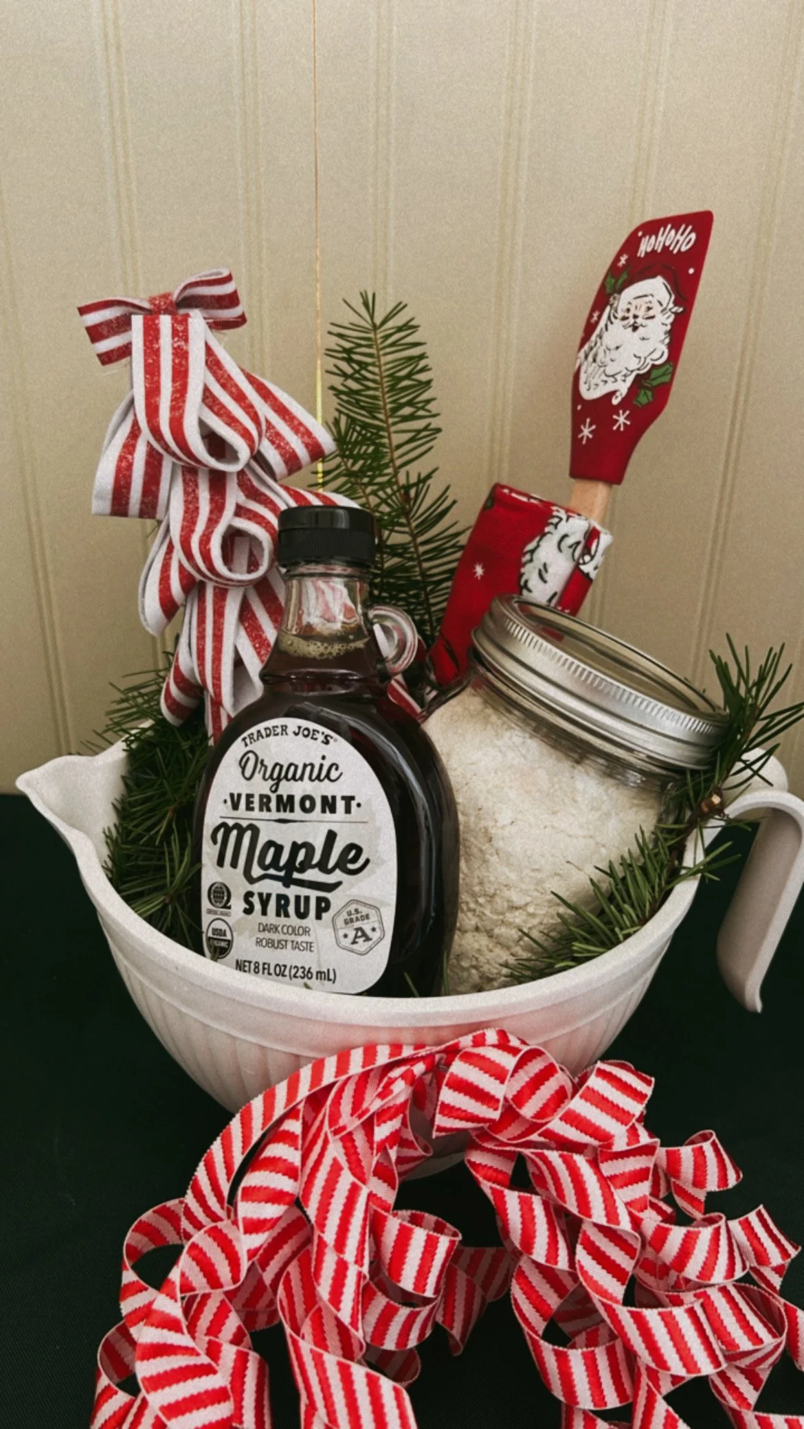 A holiday gift basket containing organic Vermont maple syrup, a jar of flour, a Santa-themed spatula, holiday ribbons, and decorative pine branches.