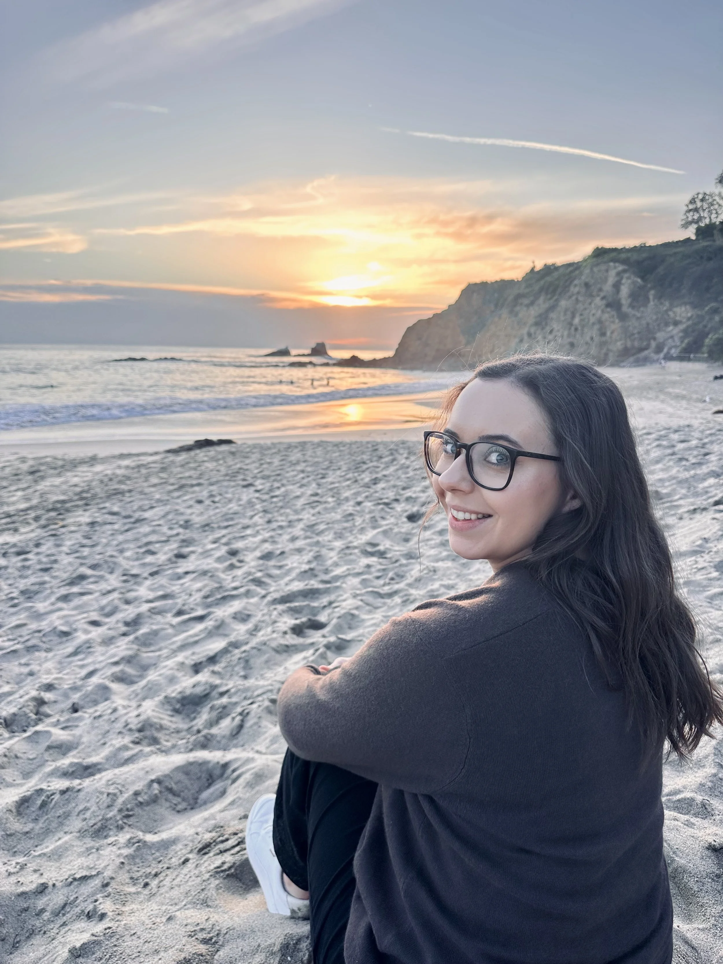 Woman smiling with glasses while sitting on the beach at sunset, wearing a brown sweater and jeans.