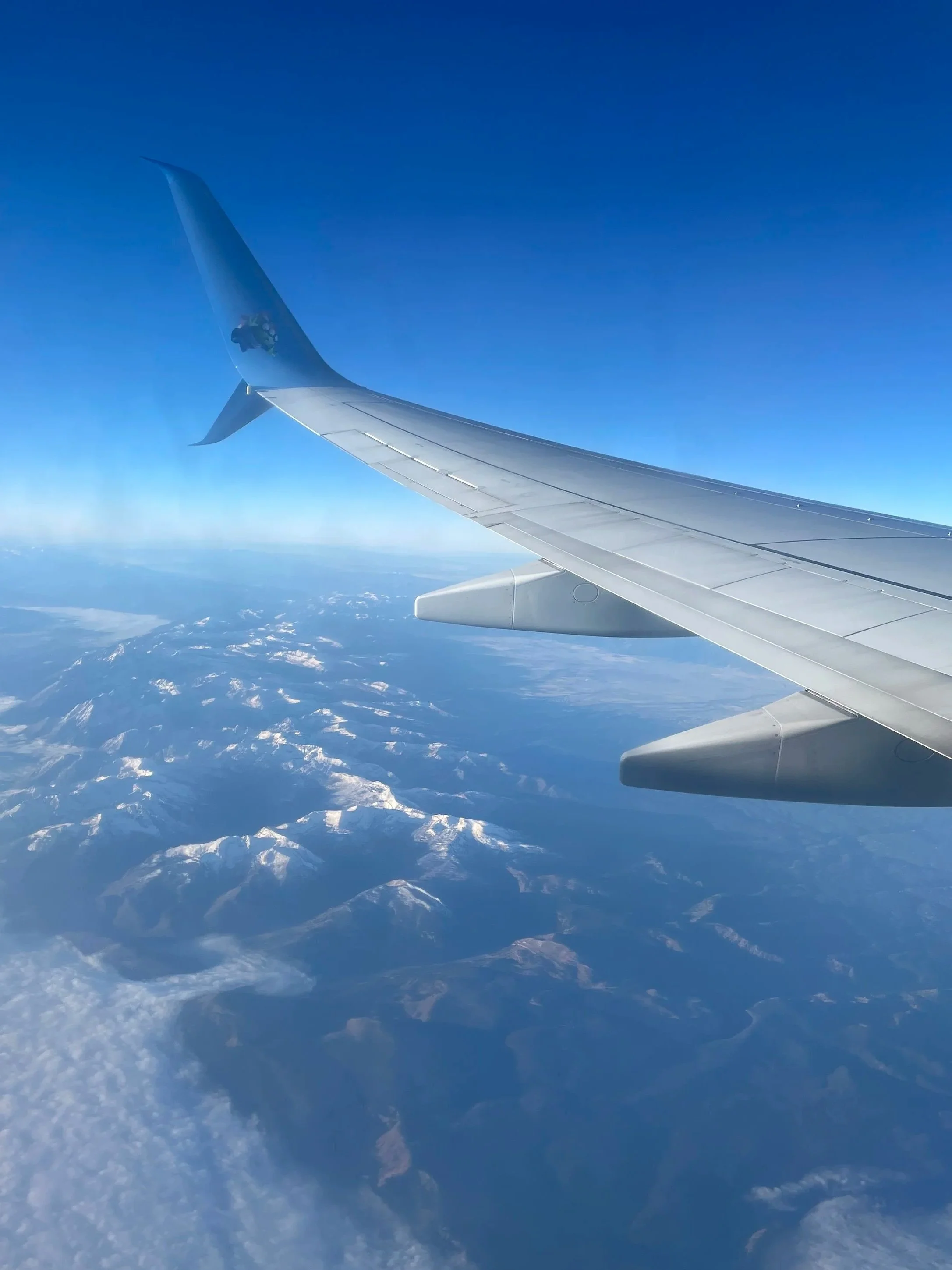 View from an airplane window showing the airplane wing and snow-capped mountains below against a clear blue sky.