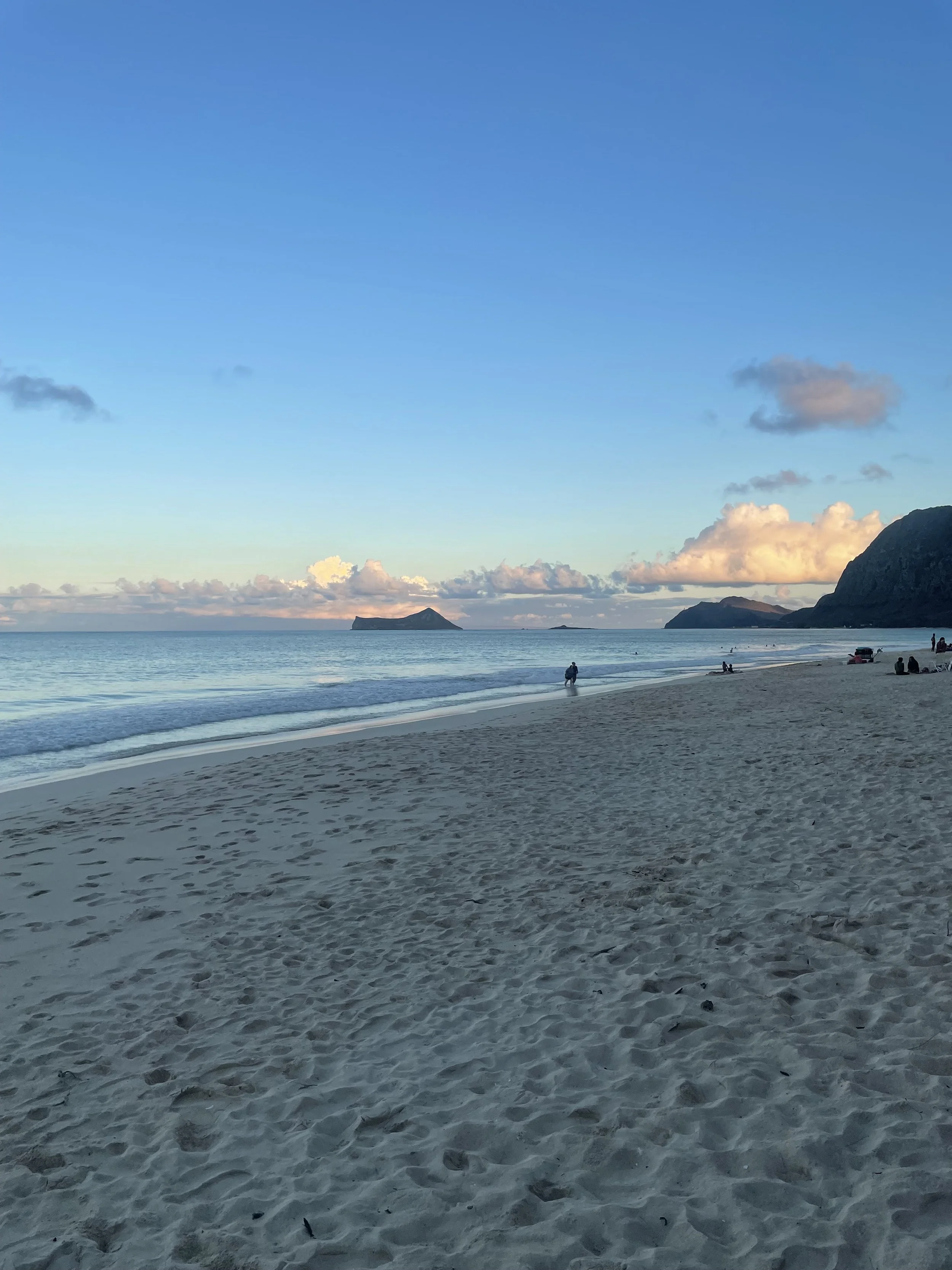A peaceful beach scene with sandy shore, ocean waves, a couple of boats, and distant islands under a clear sky with a few clouds.