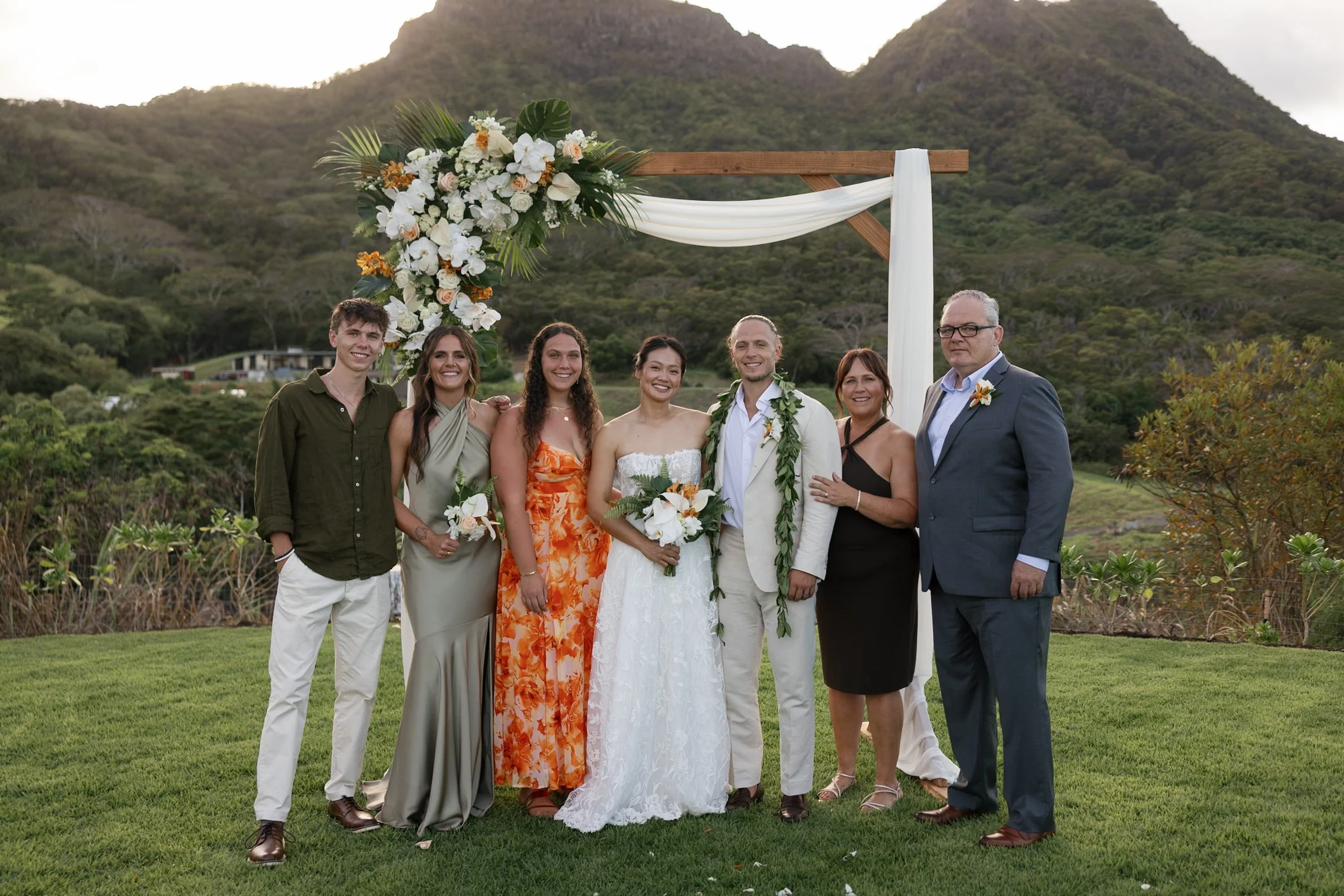 Group of seven people, including bride and groom, posing at an outdoor wedding ceremony with a mountain and greenery background. The couple stands in the center under a wooden arch decorated with flowers and fabric, surrounded by family and friends dressed in formal attire.