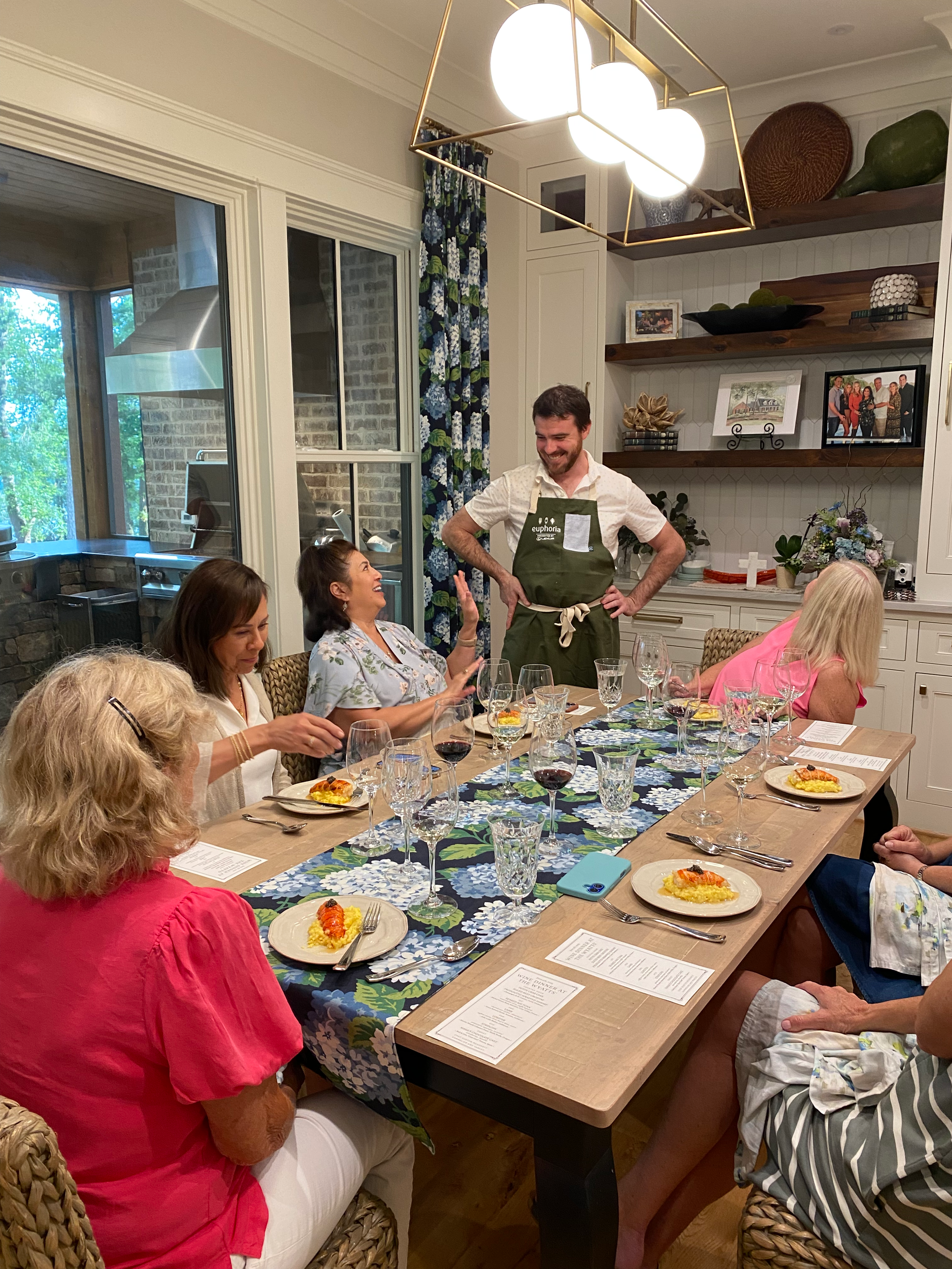 People gathered around a dining table in a well-lit, modern kitchen, preparing to enjoy a meal with wine. A man with a beard stands wearing an apron, as women sit and talk, with plates of food in front of them.