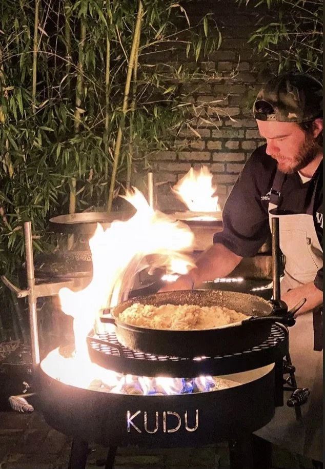 Chef Austyn McGroarty cooking over an open-flame KUDU grill at an outdoor event.