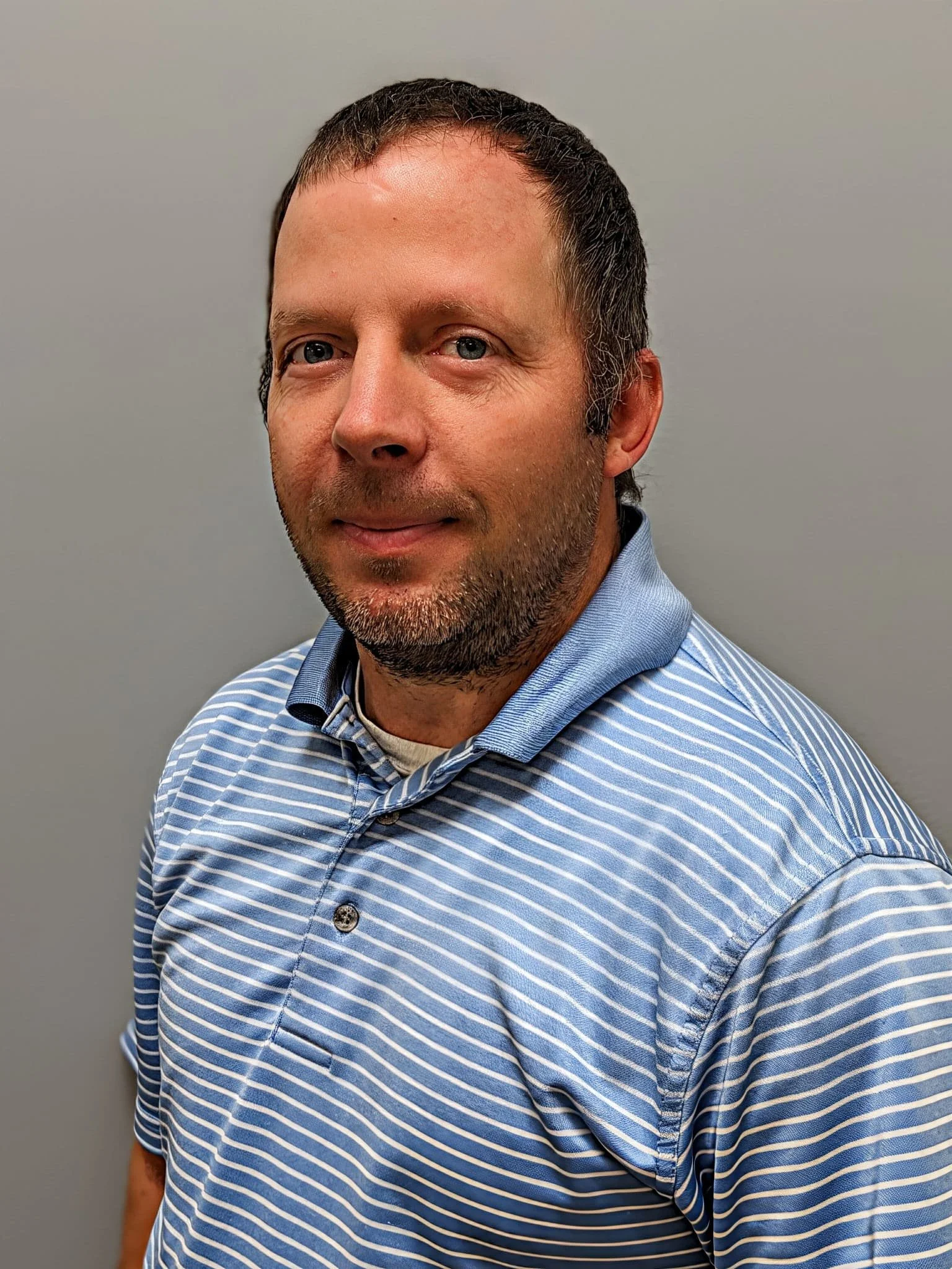 A man with short brown hair and a goatee, wearing a blue and white striped polo shirt, standing against a plain gray wall.
