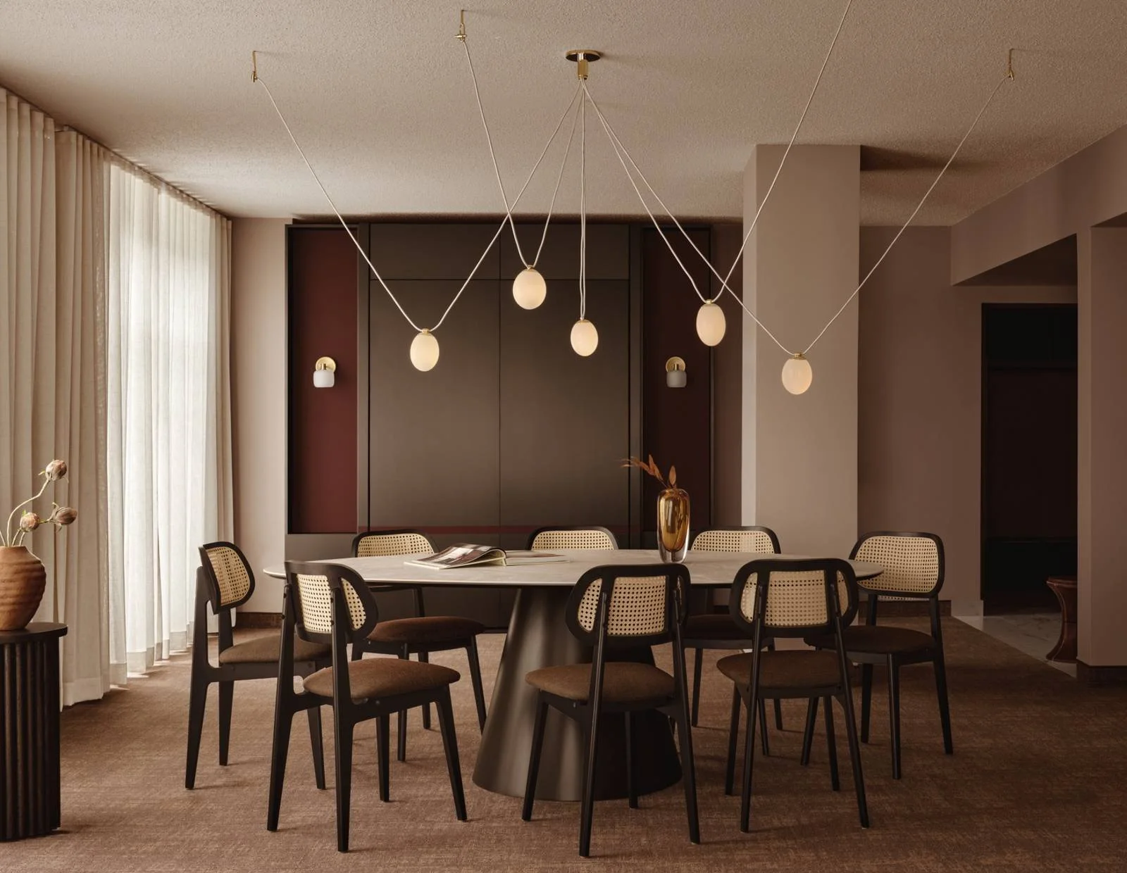 Modern dining room with round table, eight chairs, decorative pendant lights, a vase with dried flowers, and curtains by large window.