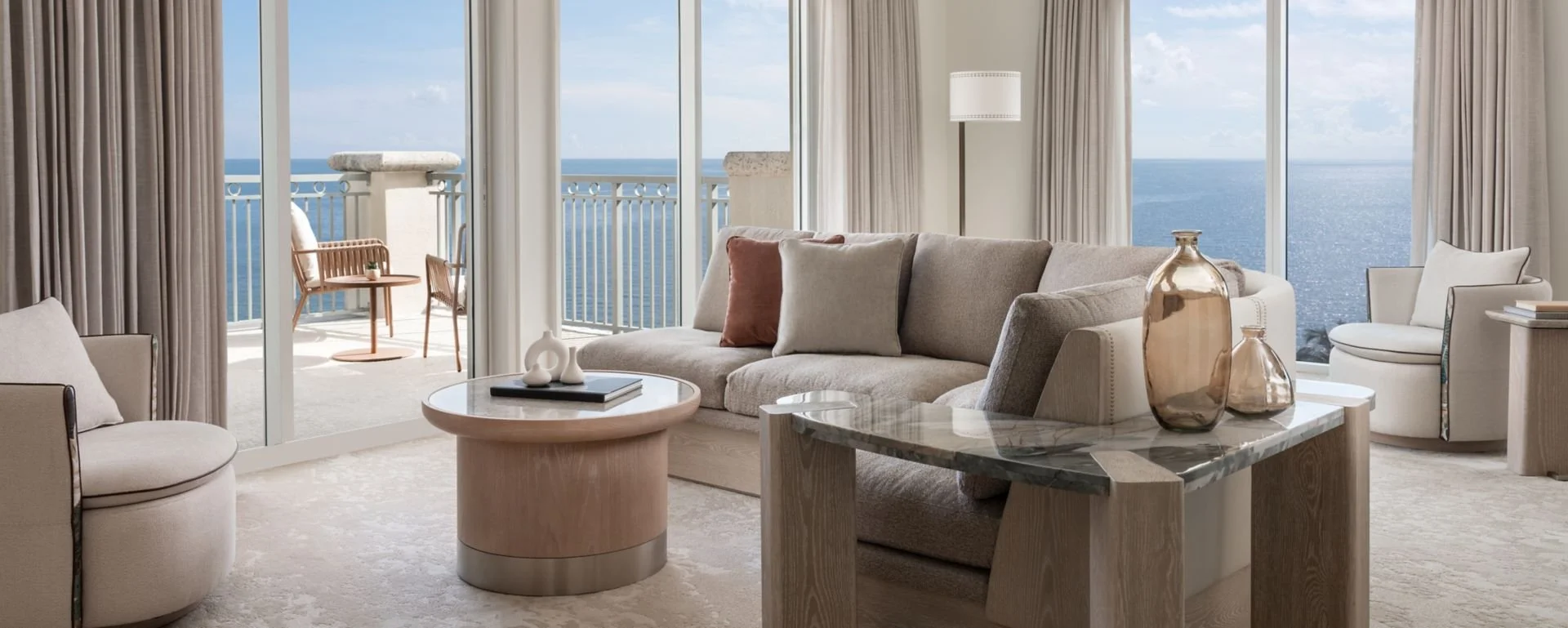 Living room with large glass sliding doors overlooking the ocean, furnished with a beige sofa, armchair, coffee table, and decorative vases, with a balcony outside.
