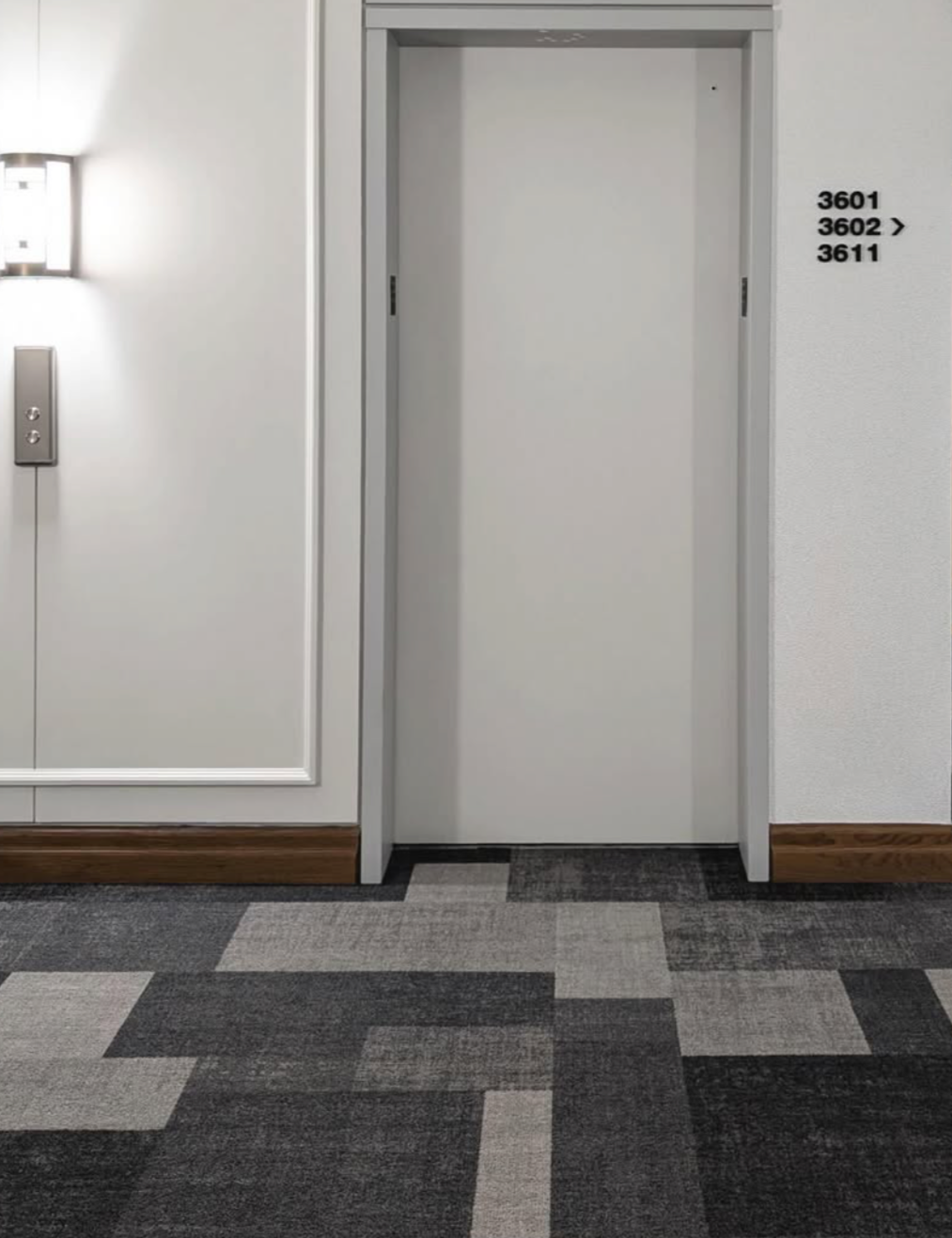An empty white elevator door next to a wall with room numbers 3601, 3602, and 3611 written on it, in a corridor with patterned carpet.