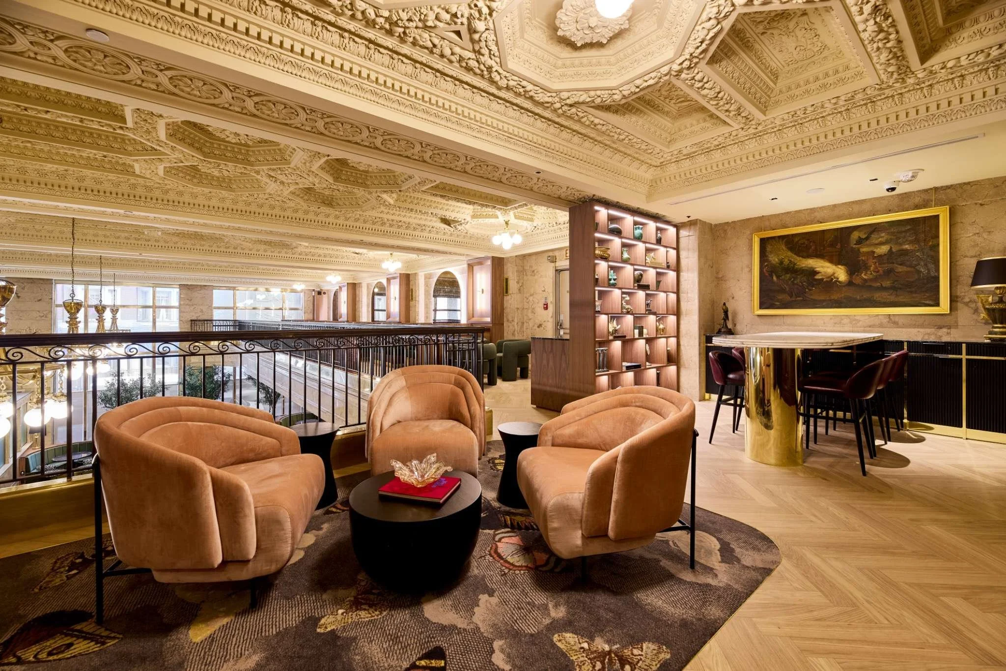 Elegant hotel lobby with ornate ceiling, cozy seating area with three peach-colored armchairs, a dark round coffee table, and a patterned rug. In the background, a wooden bookshelf, a bar with high chairs, and large windows with natural light.