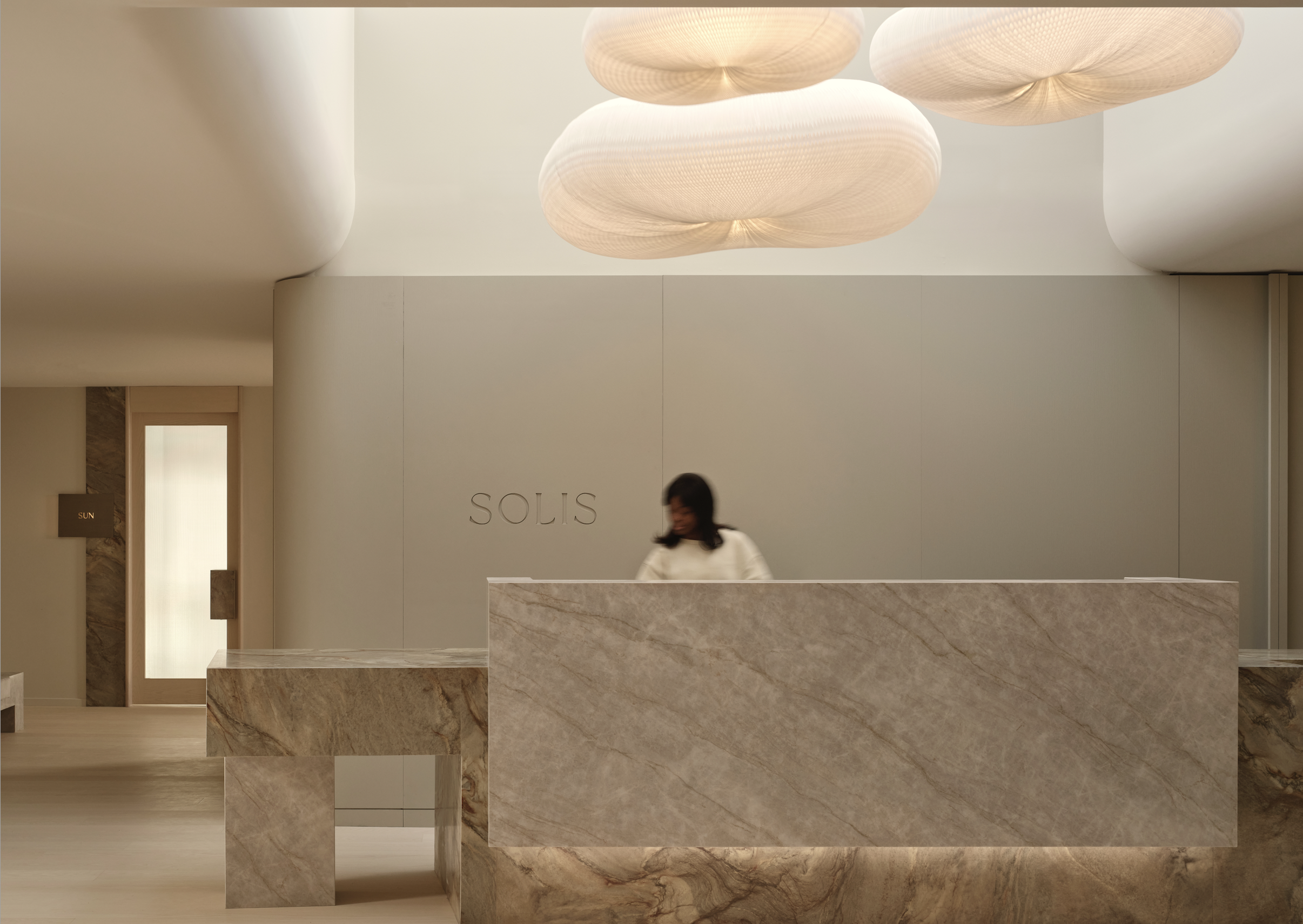 A modern hotel reception desk with a woman standing behind it, with the word 'SOLIS' on the wall behind her, decorative hanging lights above, and a door to the left labeled 'SUN'.