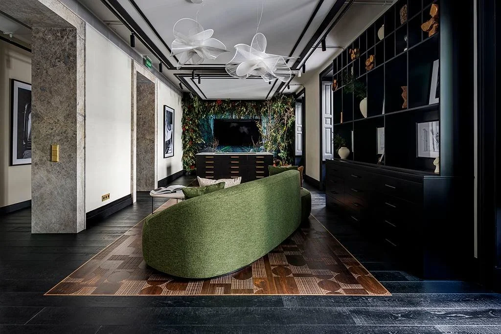 A modern living room with a green sofa, dark sustainable wood flooring, a black built-in bookshelf, framed artwork, and an abstract white ceiling light fixture.