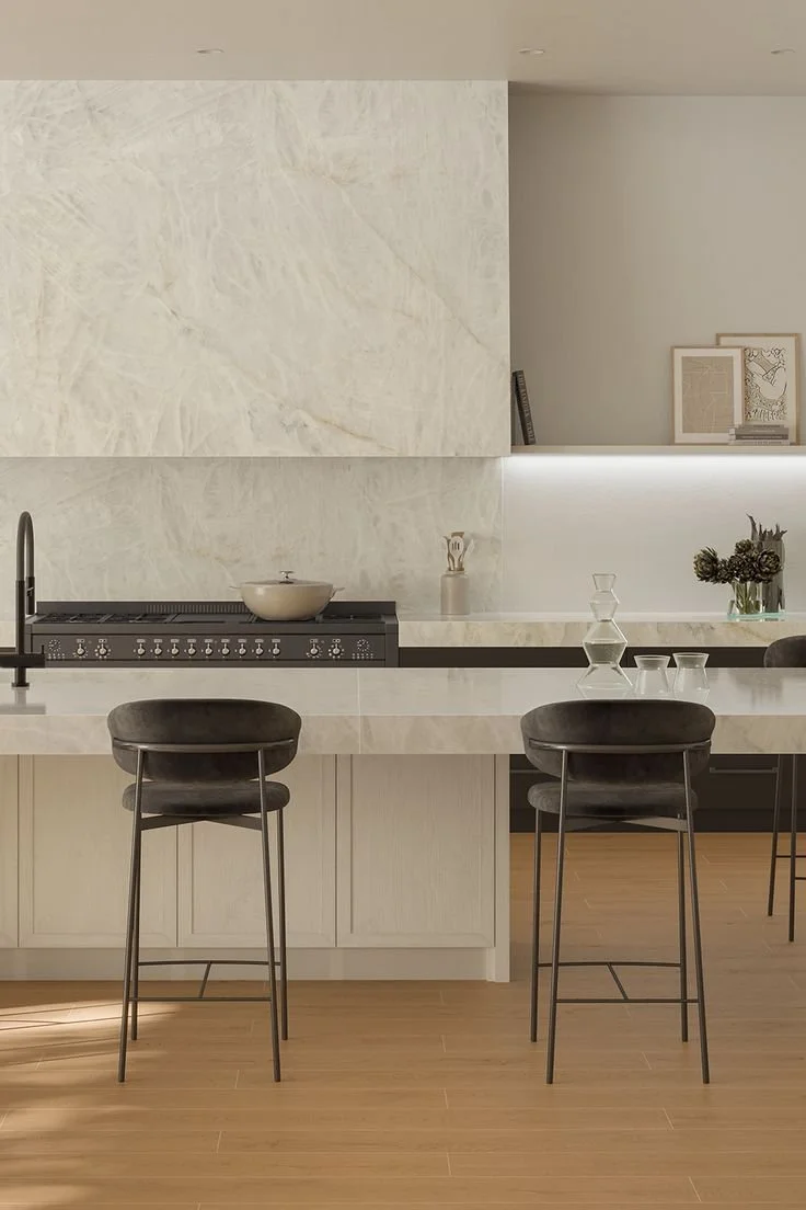 Modern kitchen with white marble countertops, black bar stools, a black stovetop, and minimalistic decor including framed art and vases and waterproof wood floors.