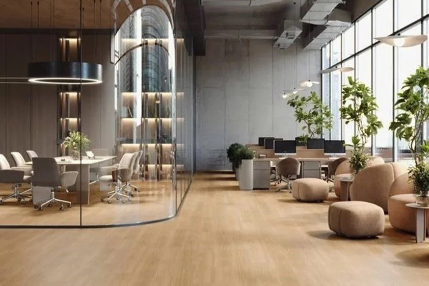 Modern office with glass-walled conference room on the left, open workspace with computers and seating on the right, and large windows with indoor plants providing natural light.