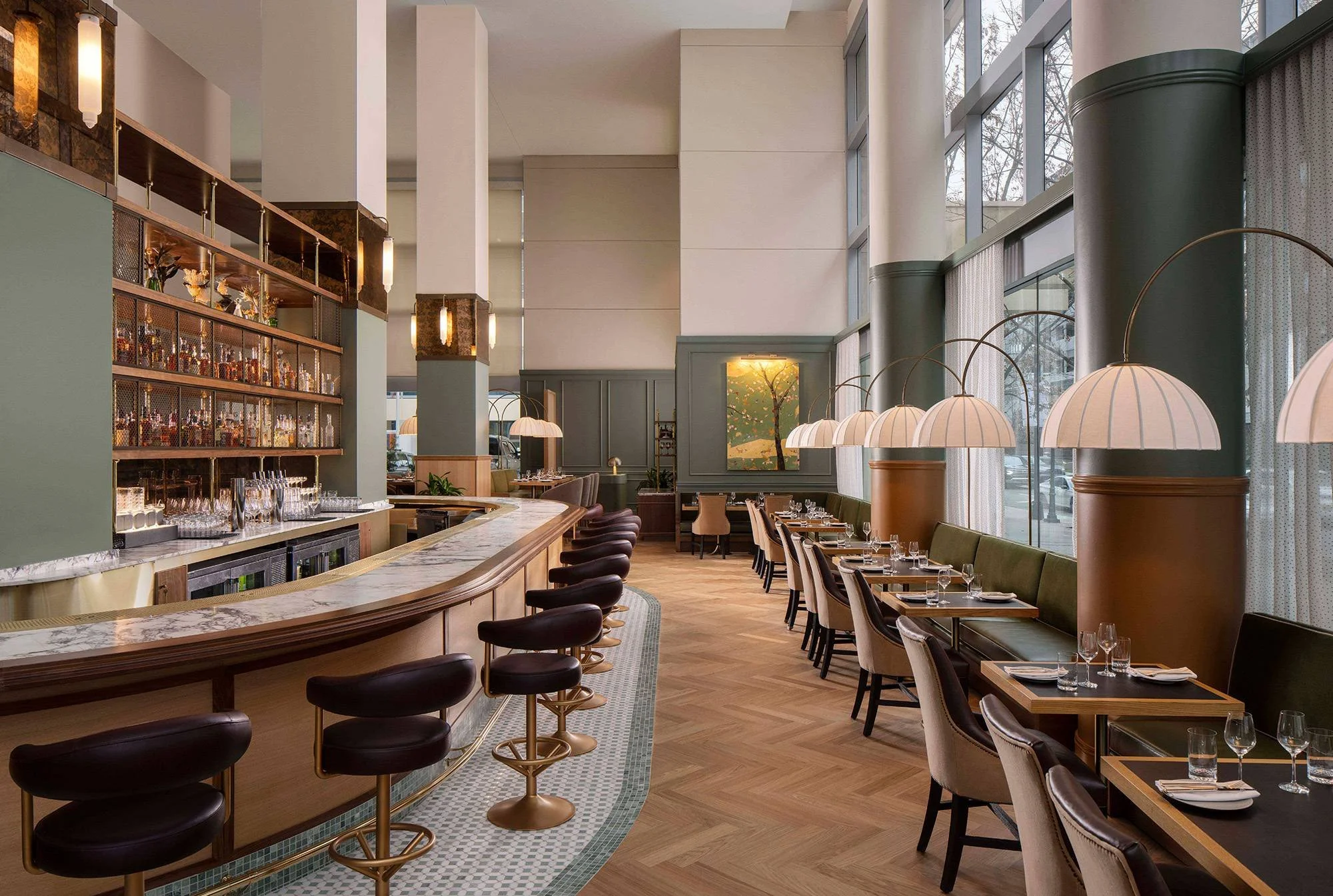 Elegant restaurant interior with a marble-topped curved bar, high black stools, and a dining area with tables set with glassware and napkins. Large windows let in natural light, with green and beige decor and hanging lamps.