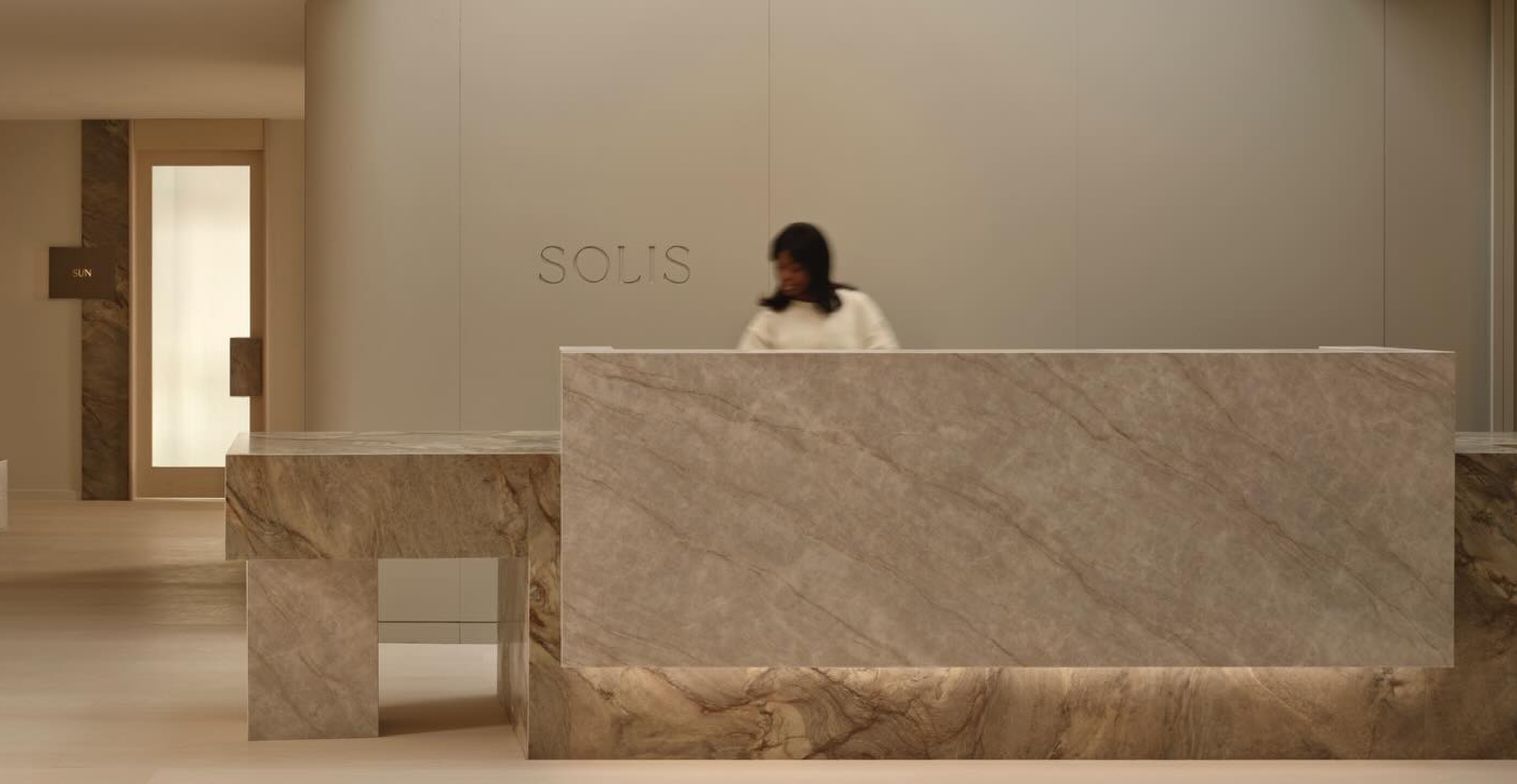A woman standing behind a marble reception desk in a modern, minimalistic lobby with a neutral color palette. The wall behind her has the word SOLIS engraved on it.