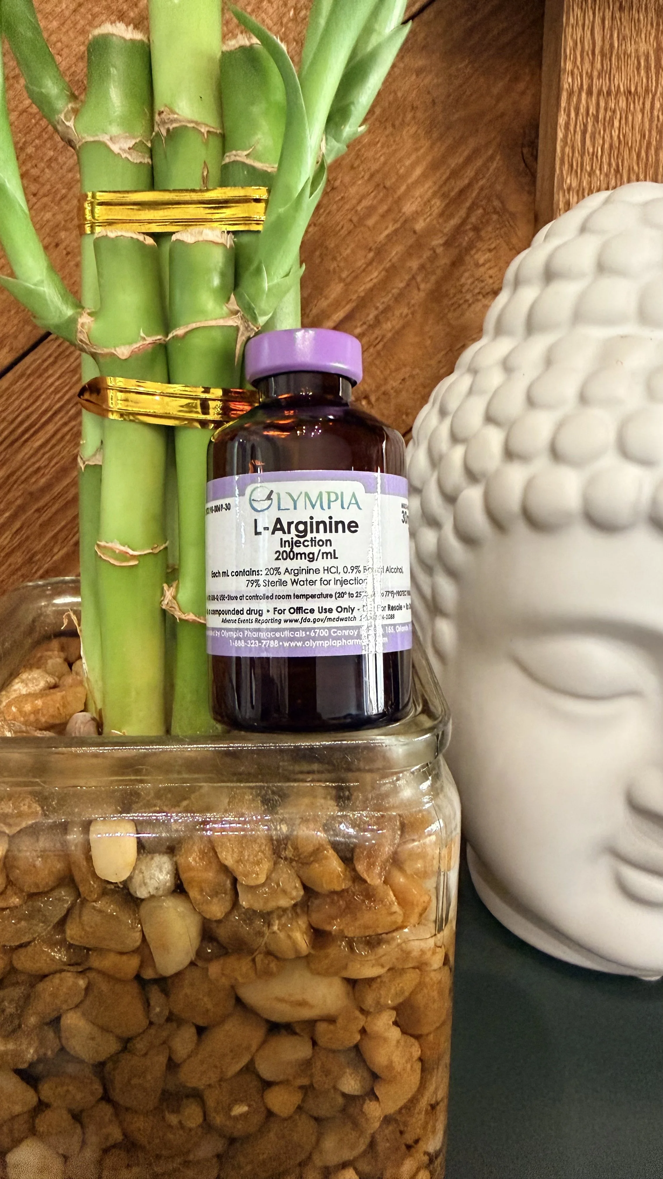 A bottle of Olympia L-Arginine injection resting against a bamboo plant with green stalks, a white Buddha head sculpture, and a glass container filled with small rocks.