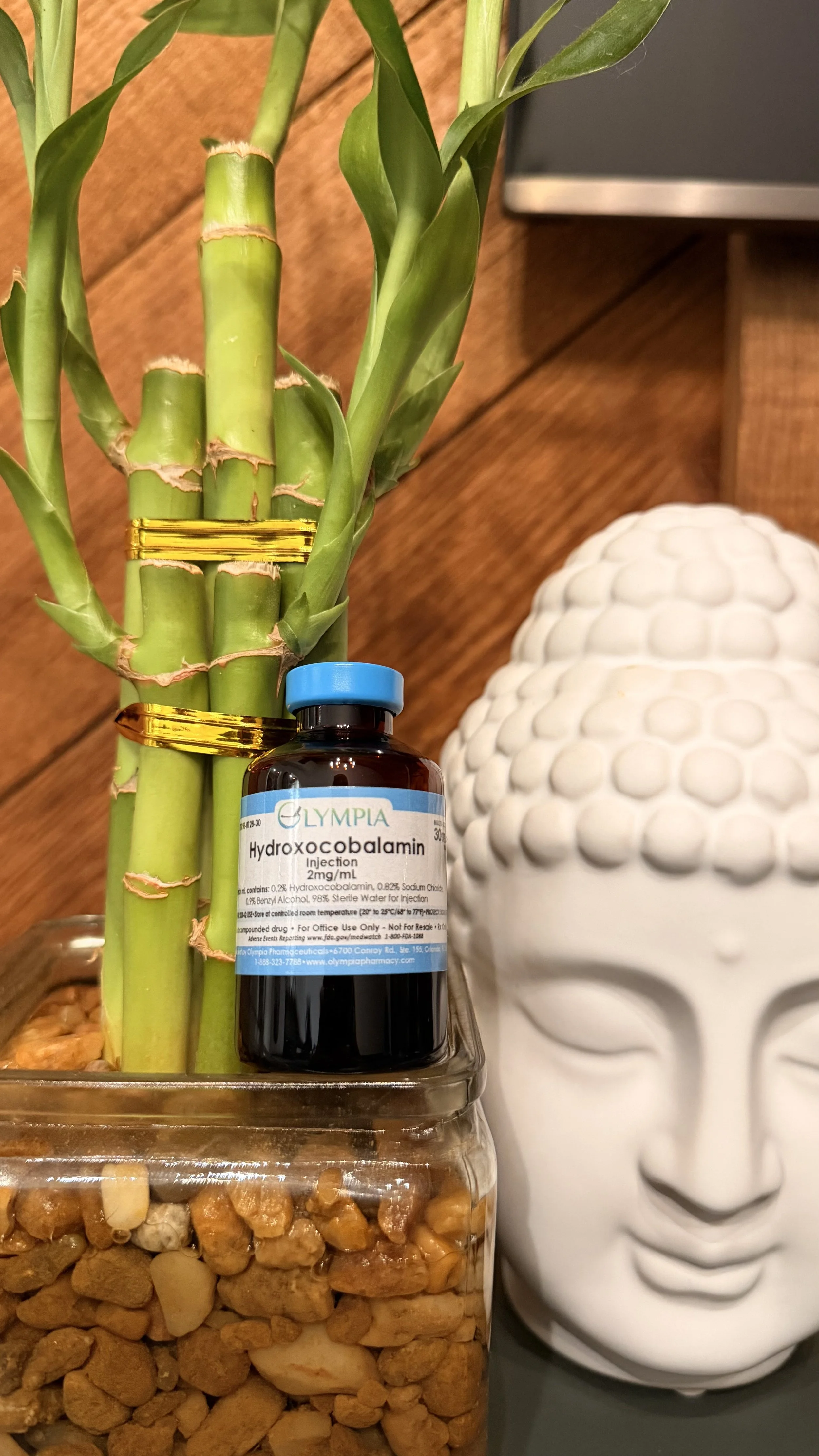 A potted bamboo plant with green stalks, a bottle of Hydroxycobalamin injection, and a white Buddha face sculpture in the background.