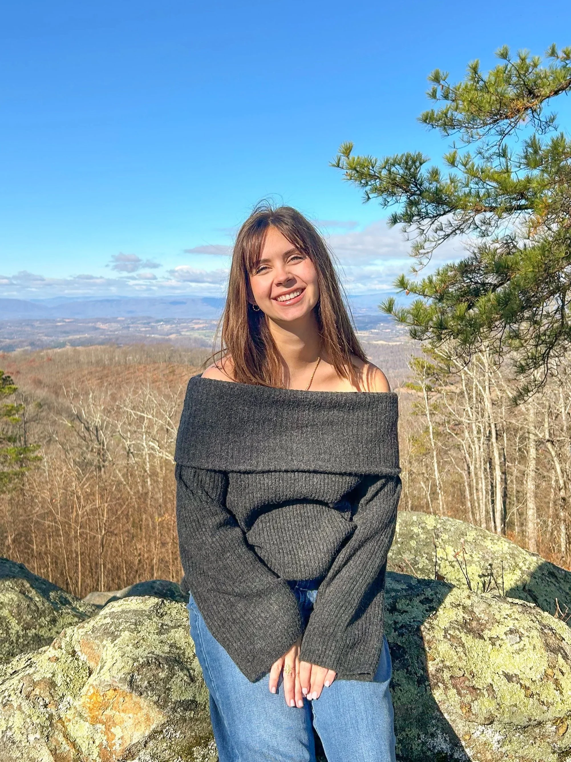 A smiling woman standing outdoors on a rocky ledge with a scenic view of trees and mountains under a clear blue sky.