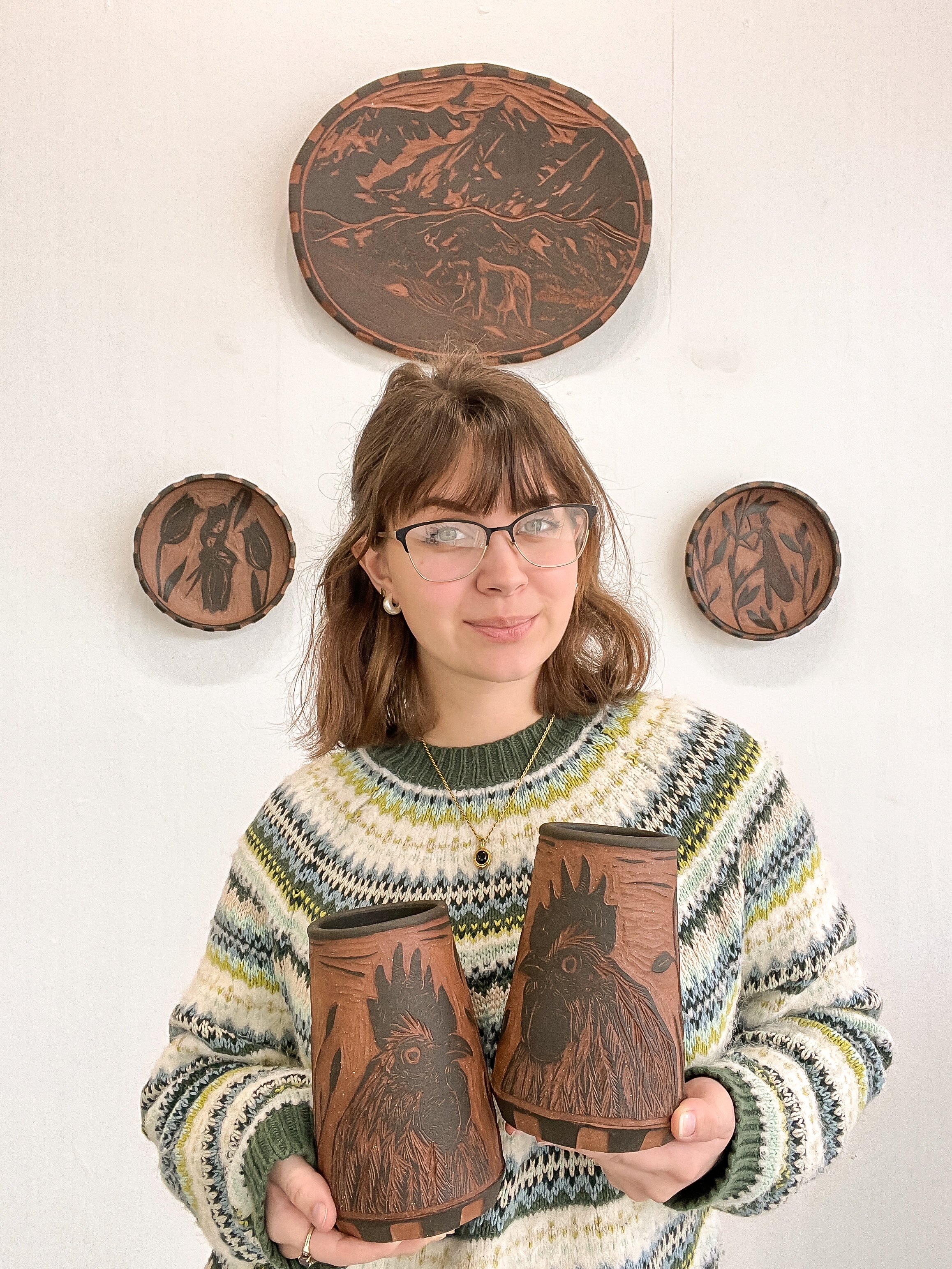 A woman with glasses and shoulder-length brown hair wearing a multicolored knit sweater is holding two carved wooden mugs with animal designs, standing in front of a white wall with three circular wood carvings featuring mountain, bird, and plant scenes.