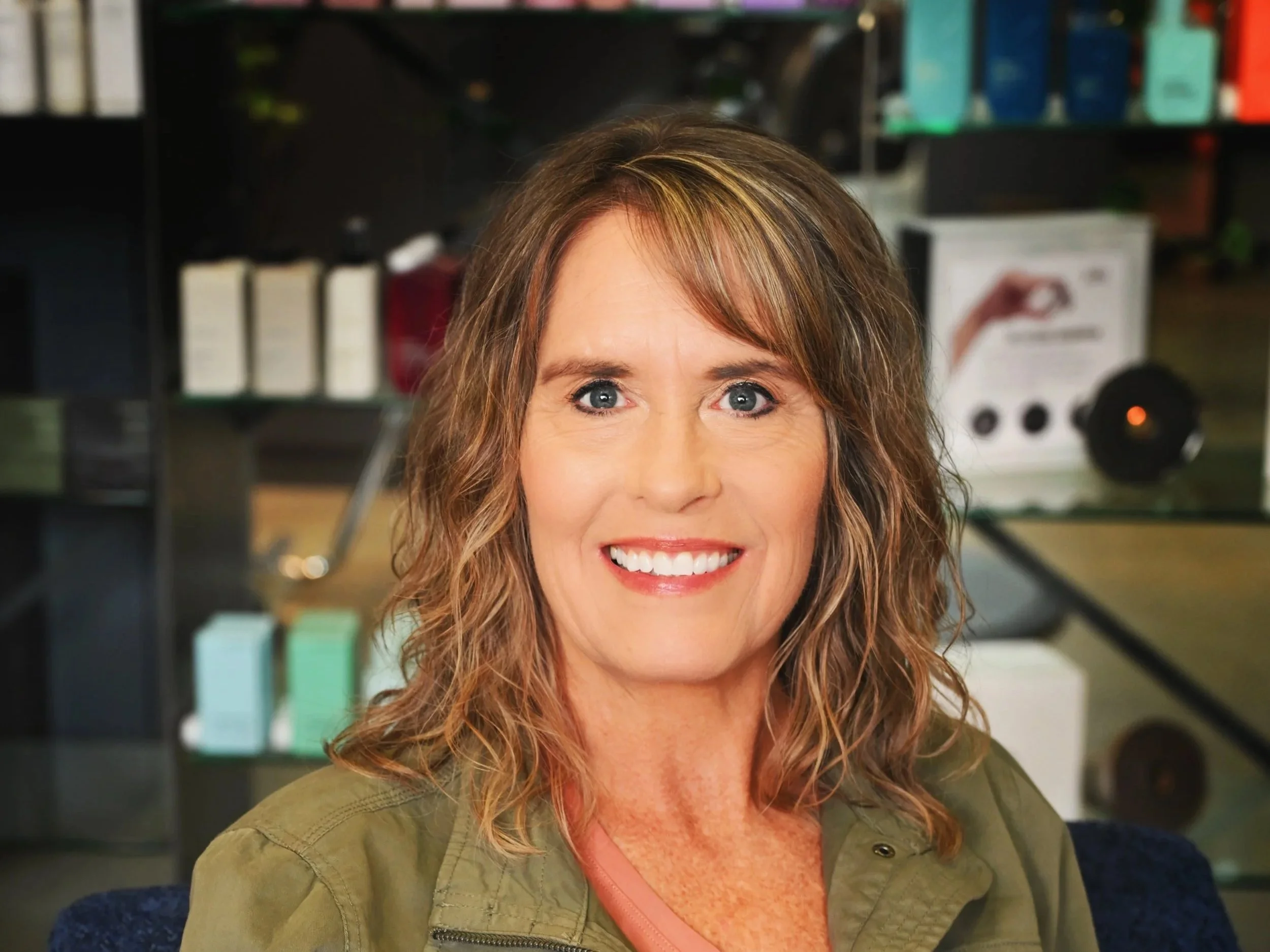 A woman with shoulder-length wavy brown hair and blue eyes smiling at the camera, wearing a green jacket inside a store or office with shelves of boxed products in the background.