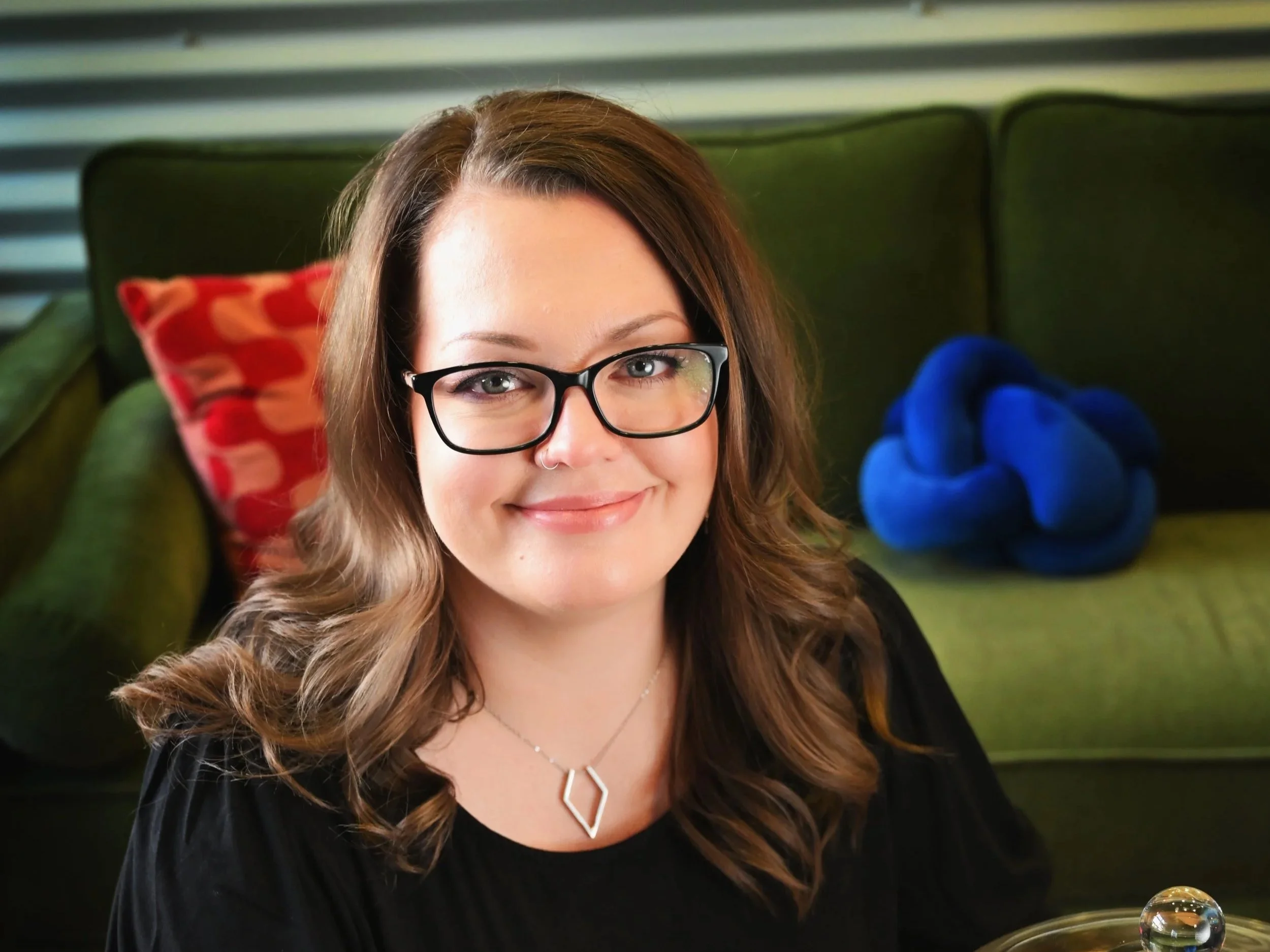 Woman with brown hair and glasses smiling, sitting on a green couch with a red patterned pillow and a blue knot sculpture in the background.