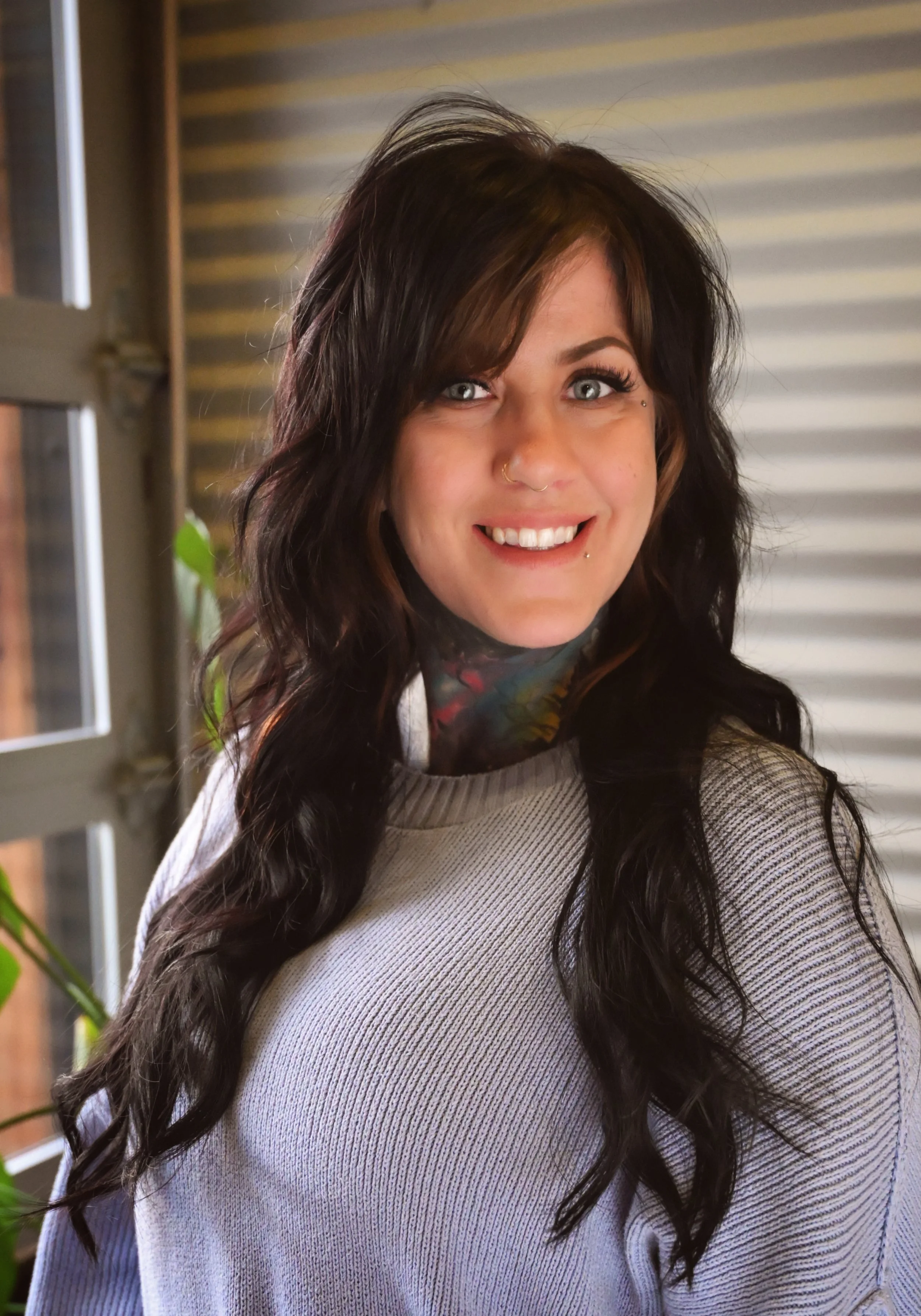 A woman with long, wavy dark hair with lighter highlights, wearing a light-colored sweater, smiling indoors near a window, with horizontal blinds and a plant nearby.