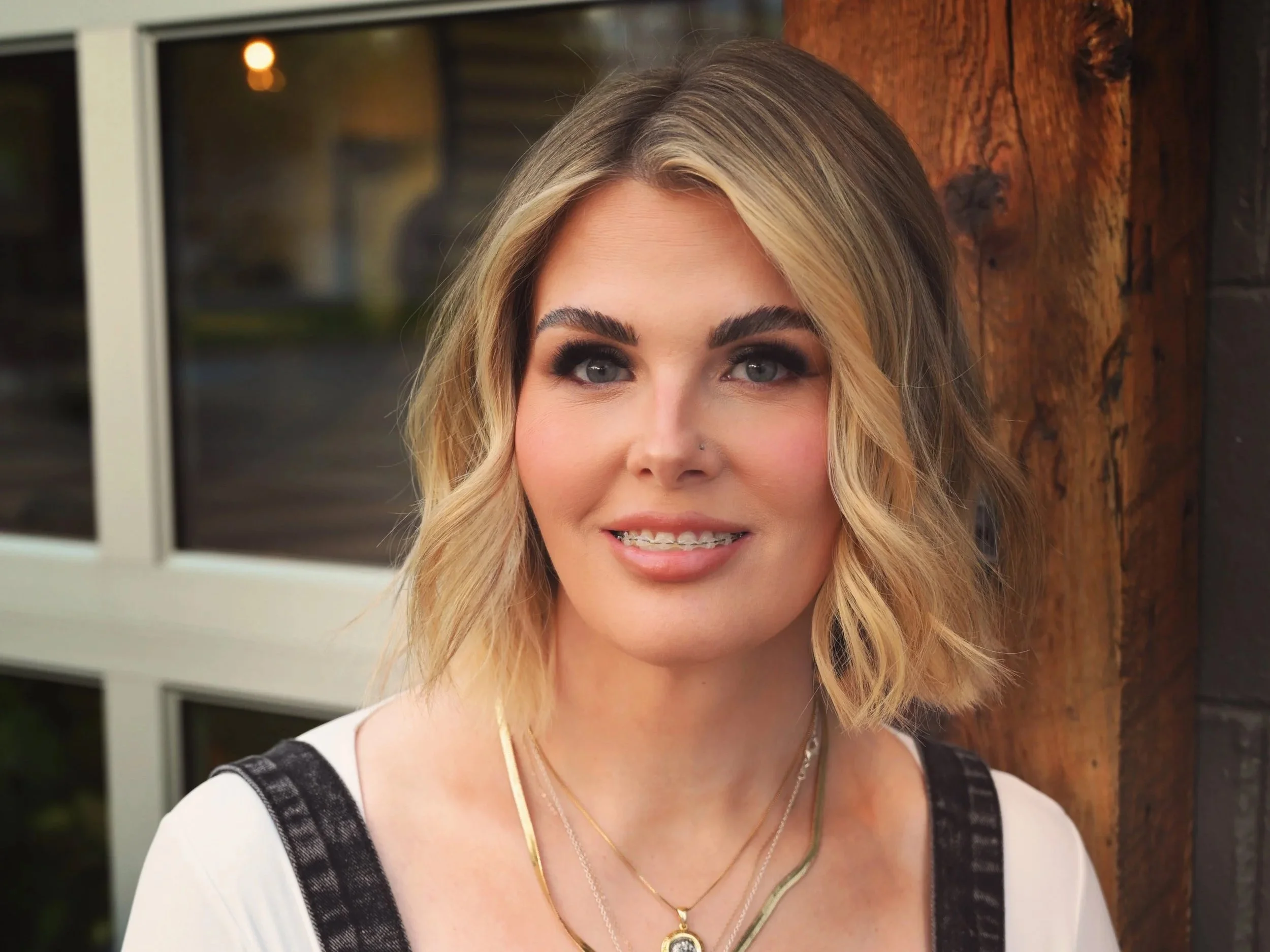 A woman with blonde hair, makeup, and jewelry smiling outdoors near a wooden wall and windows.