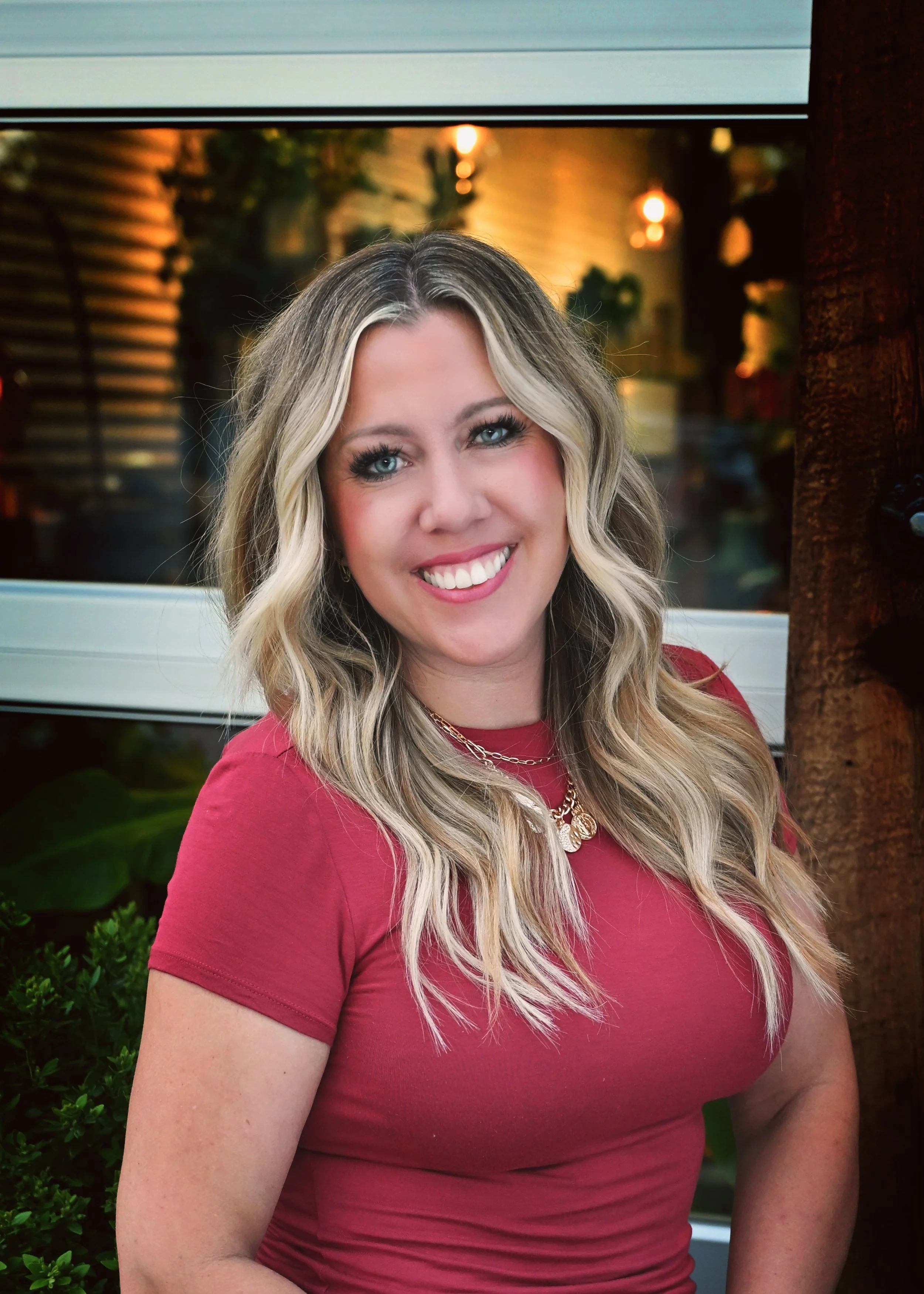 Portrait of a smiling woman with wavy blonde hair wearing a red top and layered necklaces, standing outdoors with greenery and warm lighting in the background.