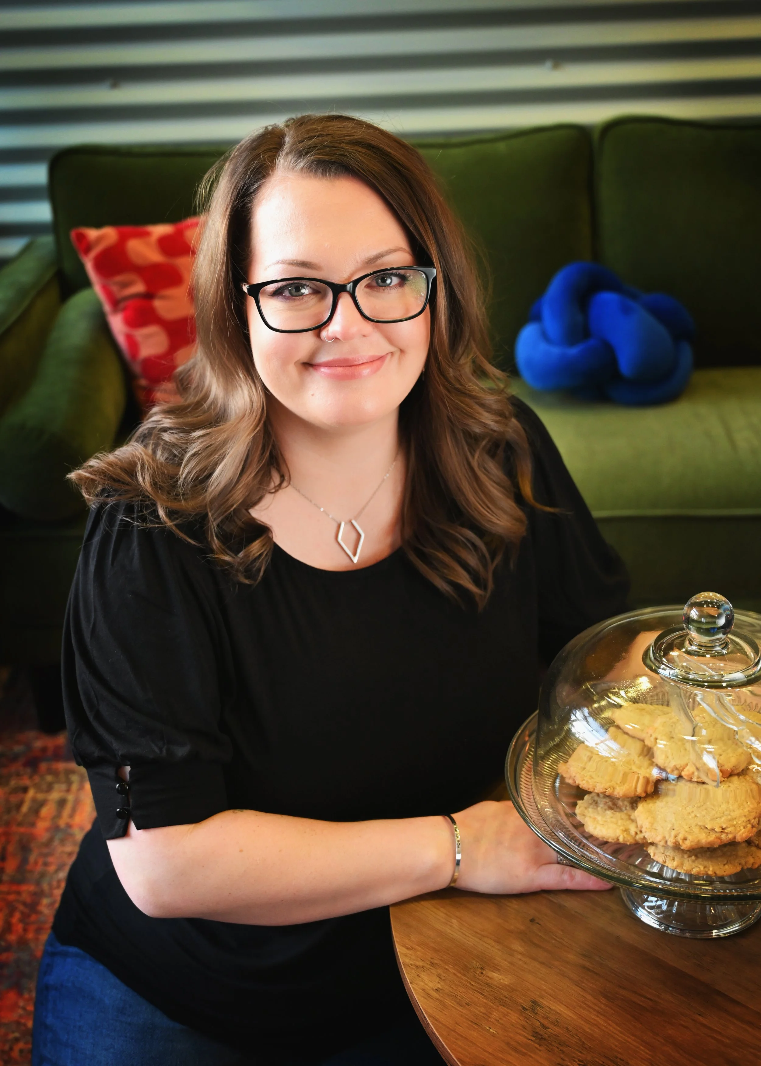 A woman with glasses and wavy brown hair smiling while holding a glass cake stand with cookies. She is sitting at a wooden table in a room with a green couch and colorful pillows in the background.